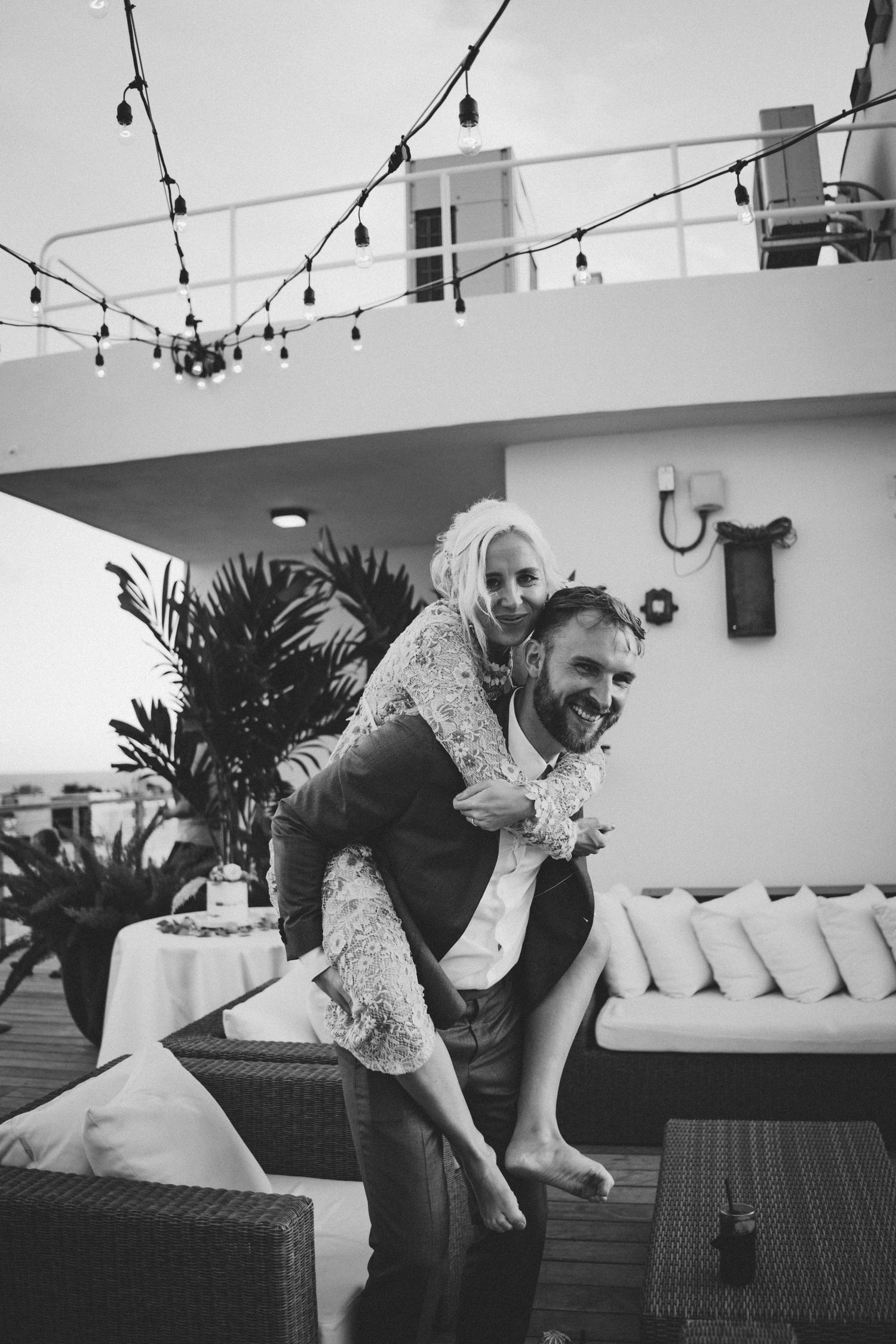 Black and white photo of a laughing groom giving his bride a piggyback ride on a rooftop terrace with string lights.