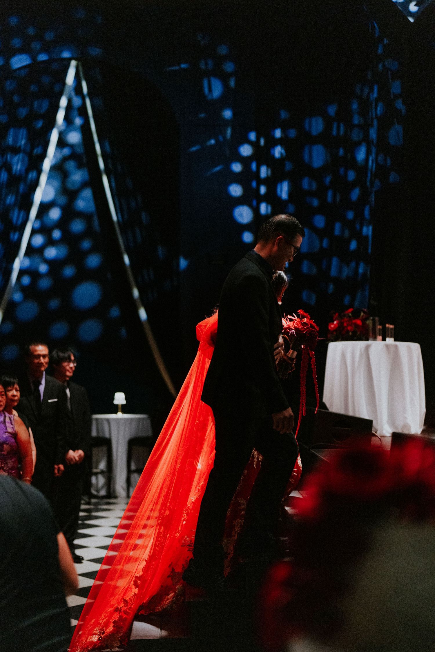 Bride in stunning red embroidered gown with long train stands with groom in black suit at altar.