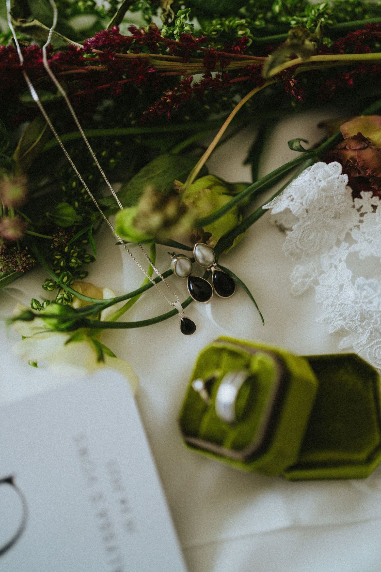 Elegant wedding flat lay with a green velvet ring box, silver earrings, floral bouquet, and lace details on white fabric.