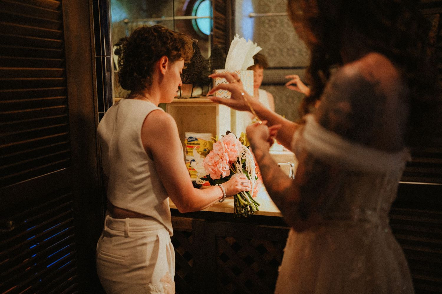 Bride and bridesmaid exchanging cards near floral bouquet in warmly lit venue before wedding ceremony.