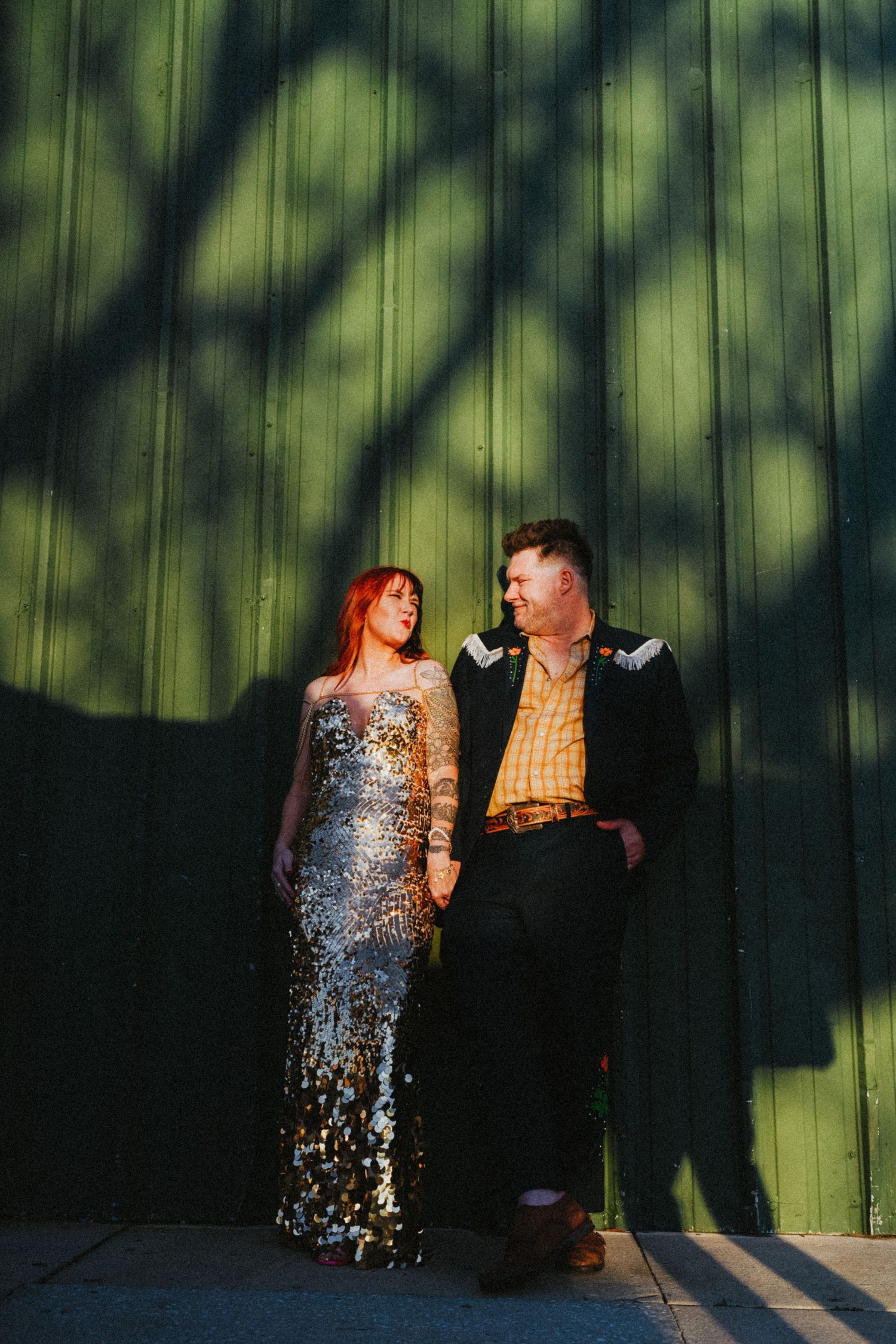 Couple in formal wear posing against a green wooden wall with dramatic tree shadows at golden hour.