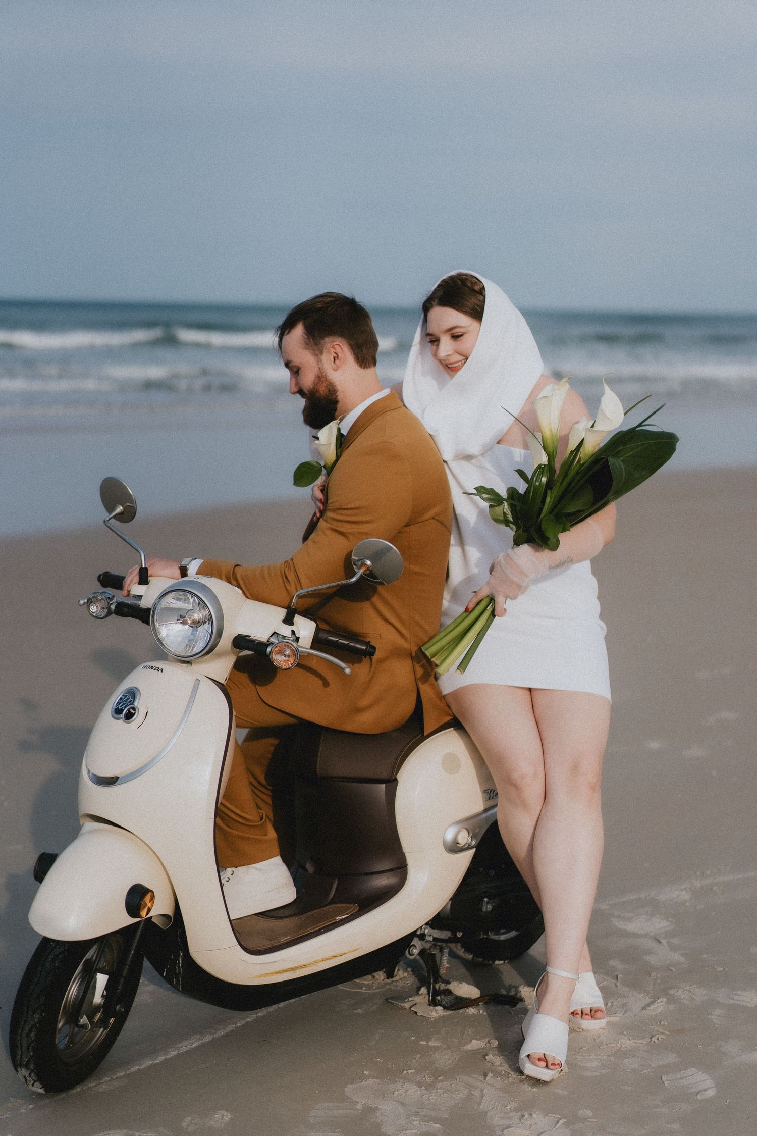 Bride and groom on vintage cream scooter at beach, bride in white mini dress holding calla lily bouquet.