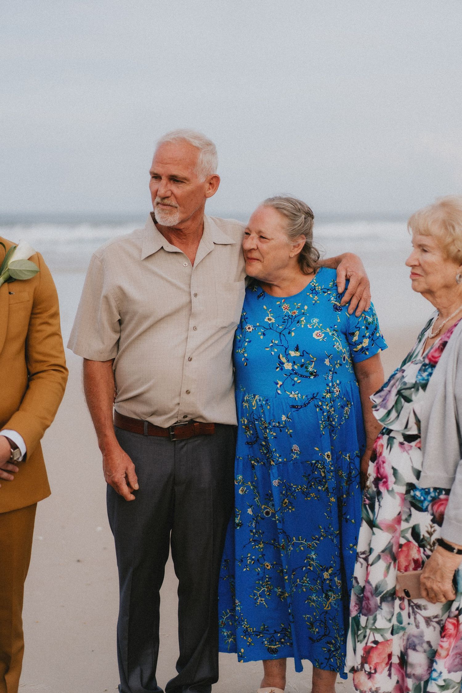 Elderly wedding guests embracing on beach, woman in blue floral dress with man in beige shirt.