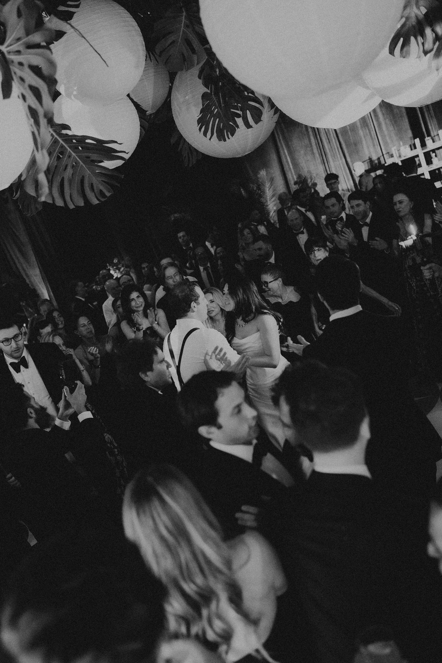 Black and white photo of elegantly dressed guests dancing at a formal wedding reception with paper lantern decorations.