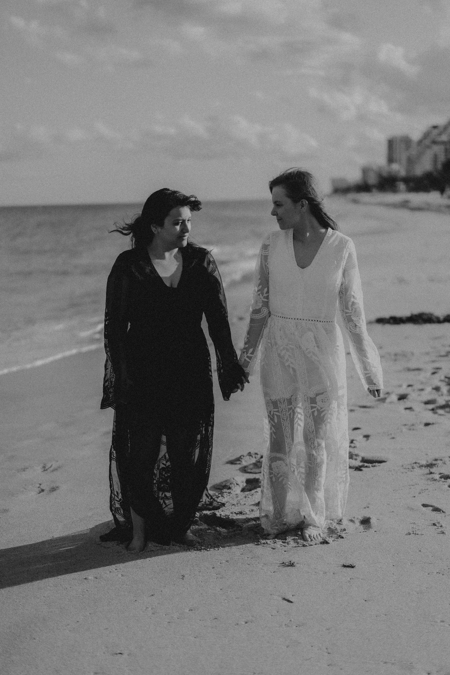Two brides holding hands walking on a beach in black and white, one in white lace gown and one in black dress.