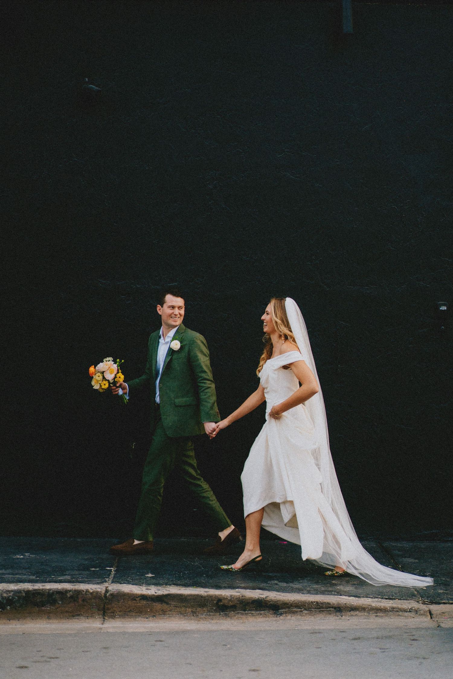 A couple walks together on a sidewalk, with the groom wearing a green suit and bride in flowing white dress.