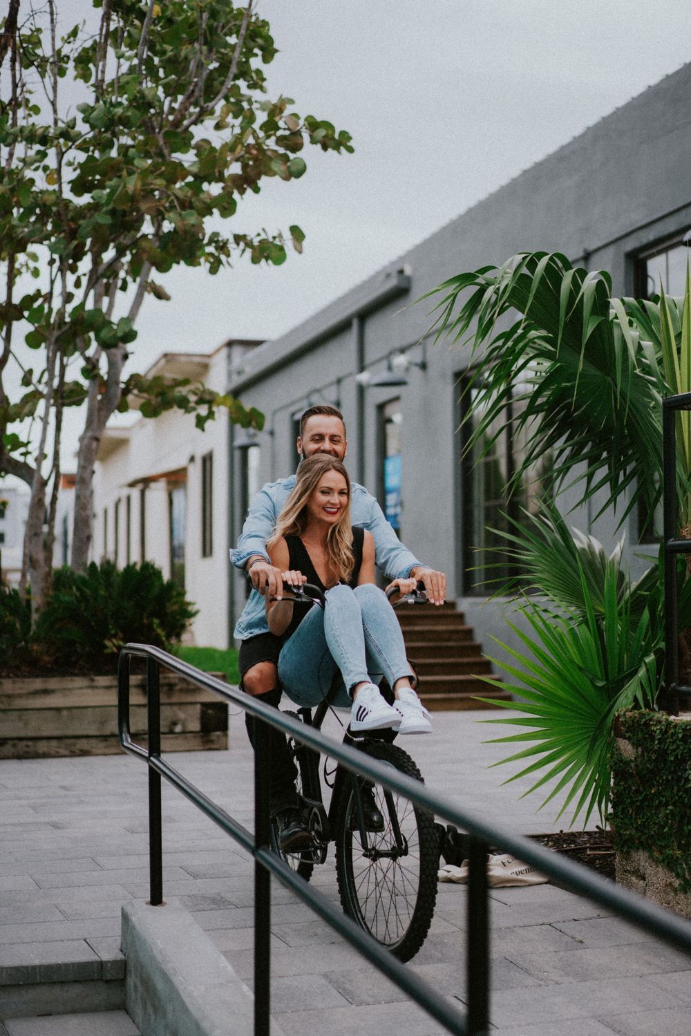 People playfully pose with a bicycle near palm trees and concrete steps.