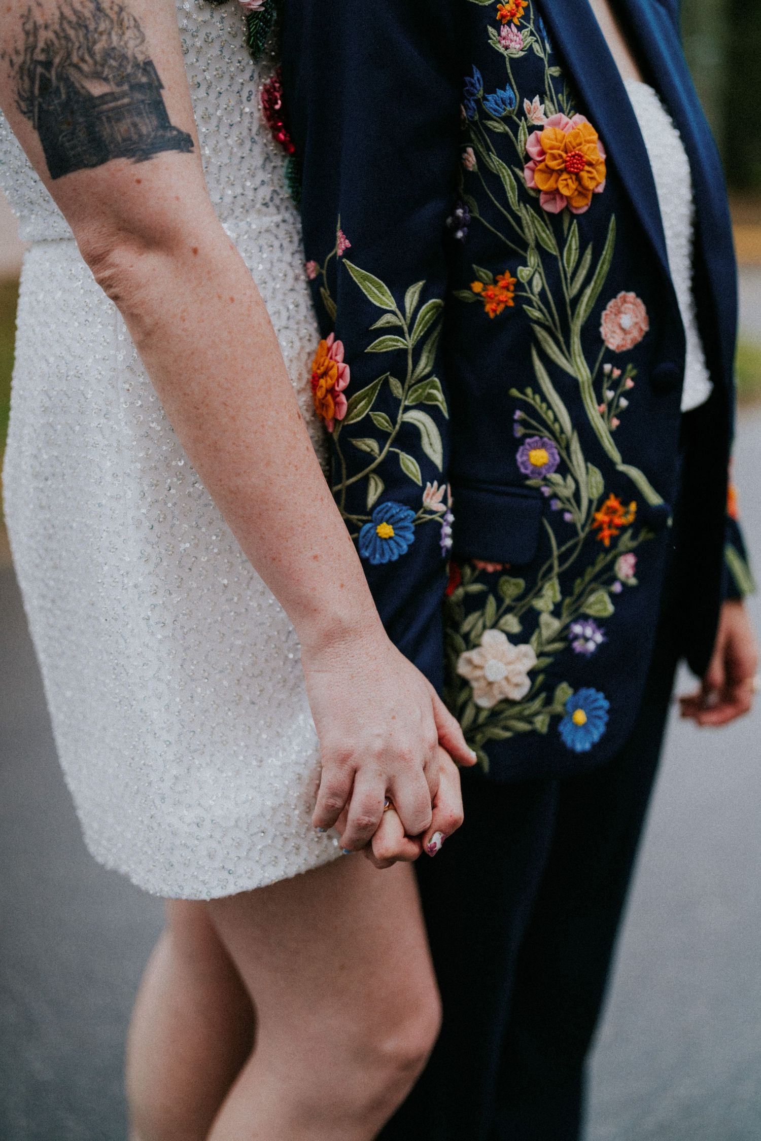 Couple holding hands, one in white dress with tattoo, other in floral embroidered navy blazer, at a wedding.