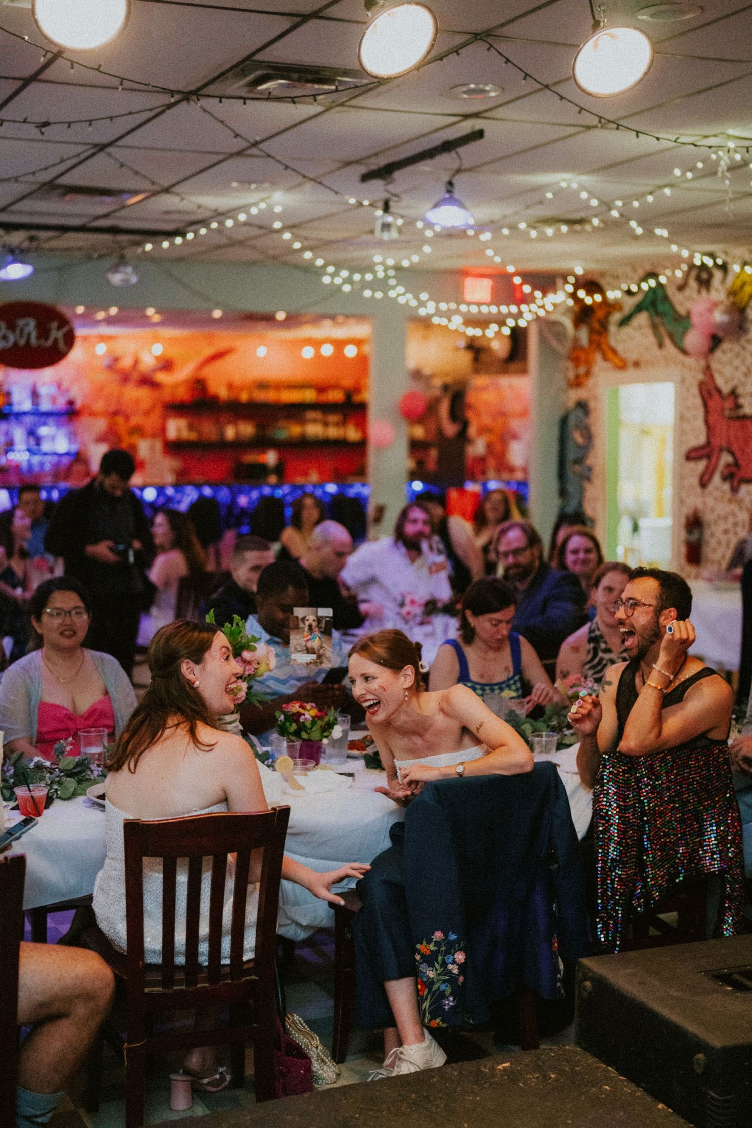 Guests laughing and celebrating at a colorful wedding reception with string lights and decorated tables.
