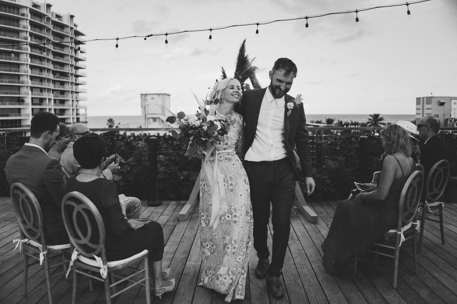 Black and white photo of a bride and groom walking hand in hand on a rooftop wedding venue with guests seated nearby.