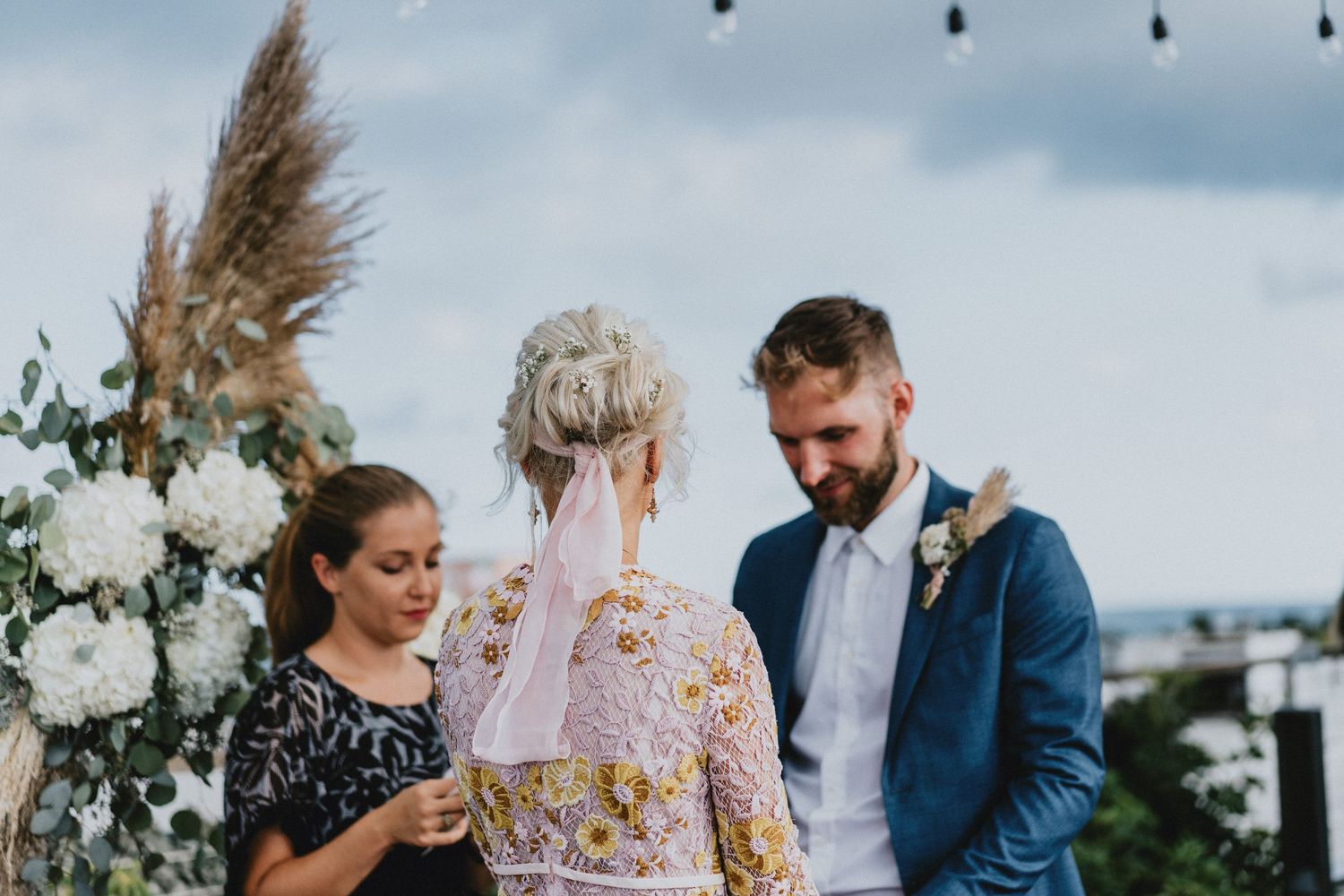 Couple exchanging vows at outdoor wedding ceremony with white floral arch and officiant on cloudy day.