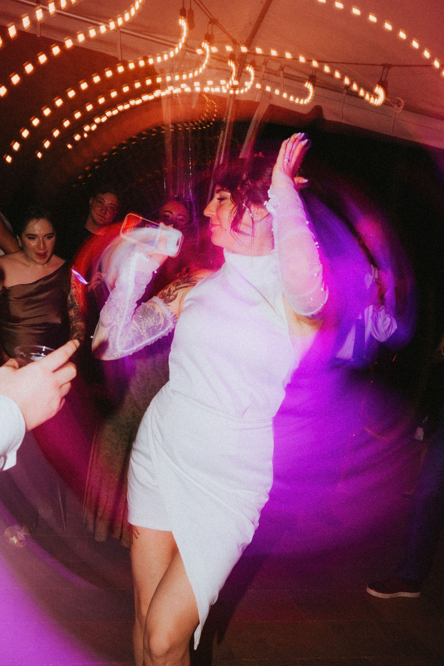 Bride in white mini dress dancing joyfully at wedding reception with purple lights and string lights overhead.