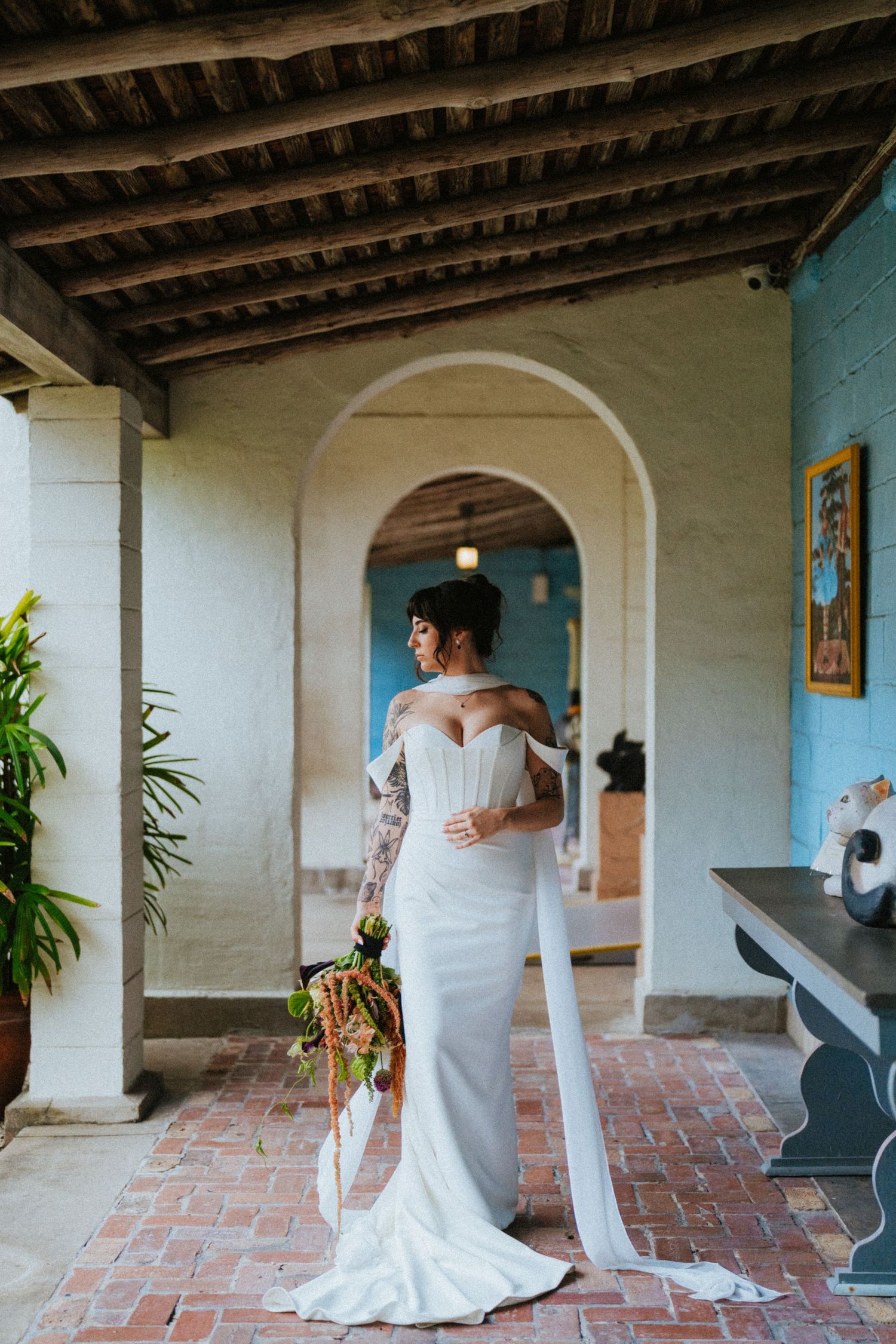 Tattooed bride in off-shoulder white gown holds bouquet in arched colonial venue with brick floors.