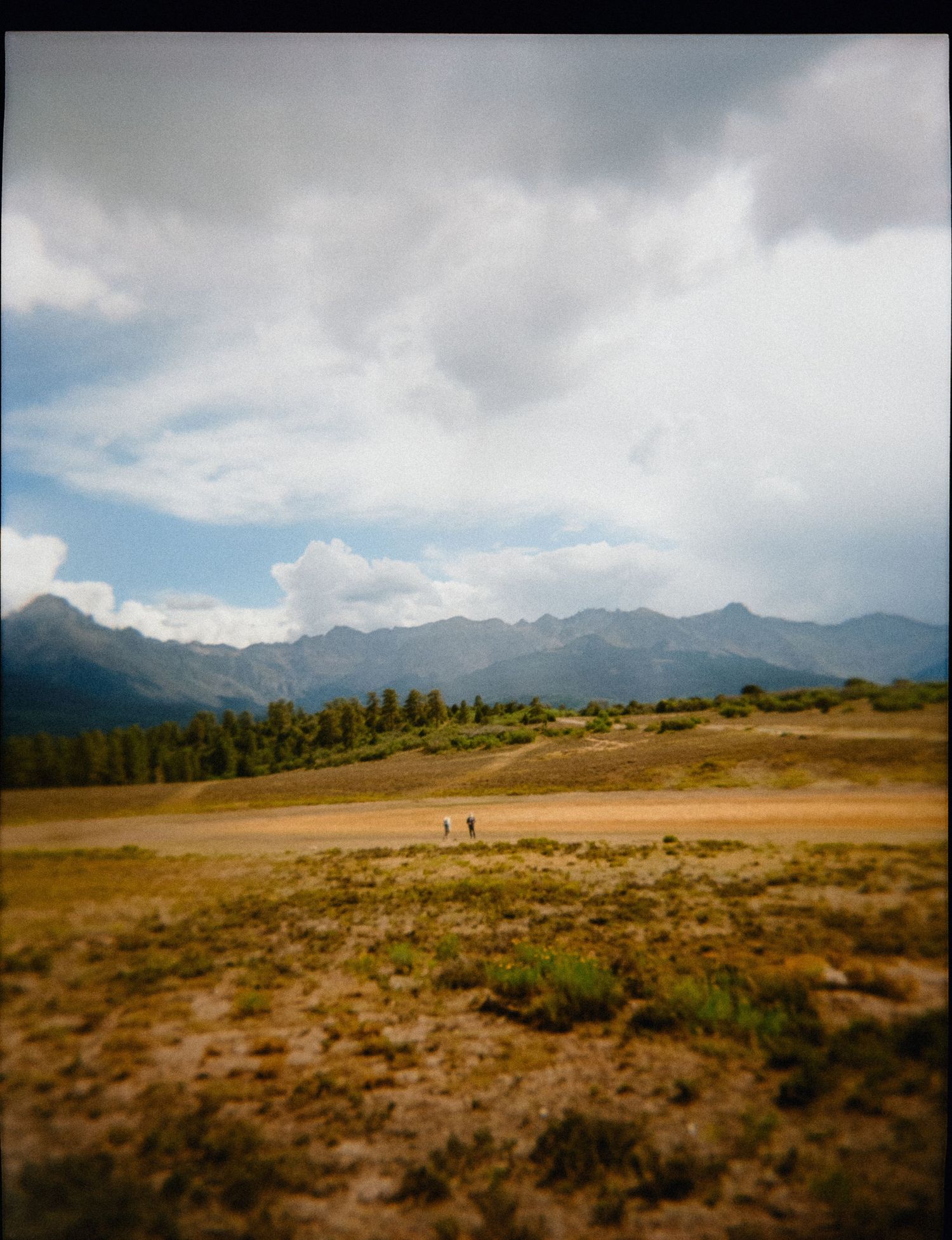 Two hikers walk across a vast open meadow with dramatic mountain peaks and stormy clouds in the background.
