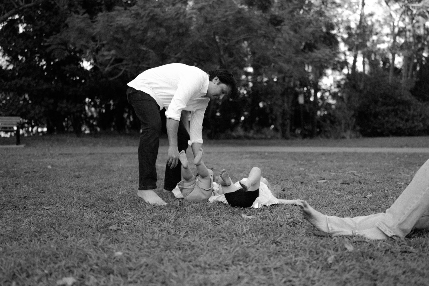 Black and white photo of a parent bending down to play with a baby on grass in a park.
