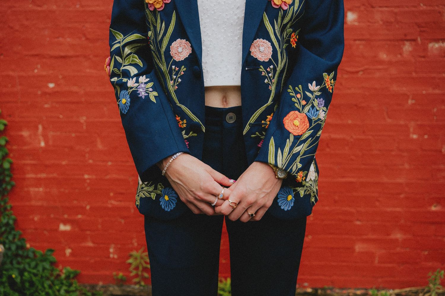 Woman wearing navy floral embroidered blazer with colorful flowers, paired with dark jeans, standing against red brick wall.