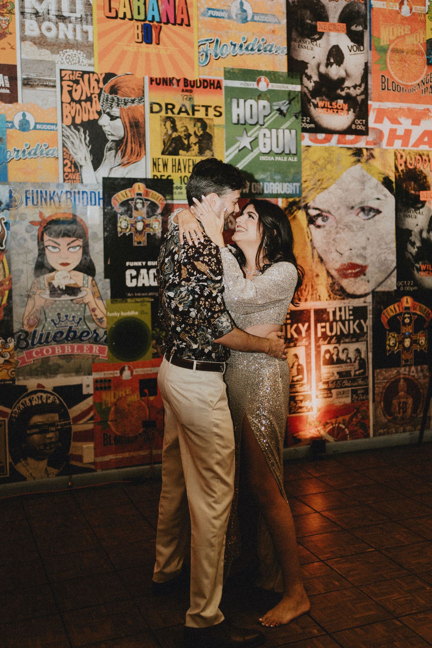 Couple shares a romantic embrace in front of a vibrant concert poster wall, dressed in floral shirt and silver sequin gown.