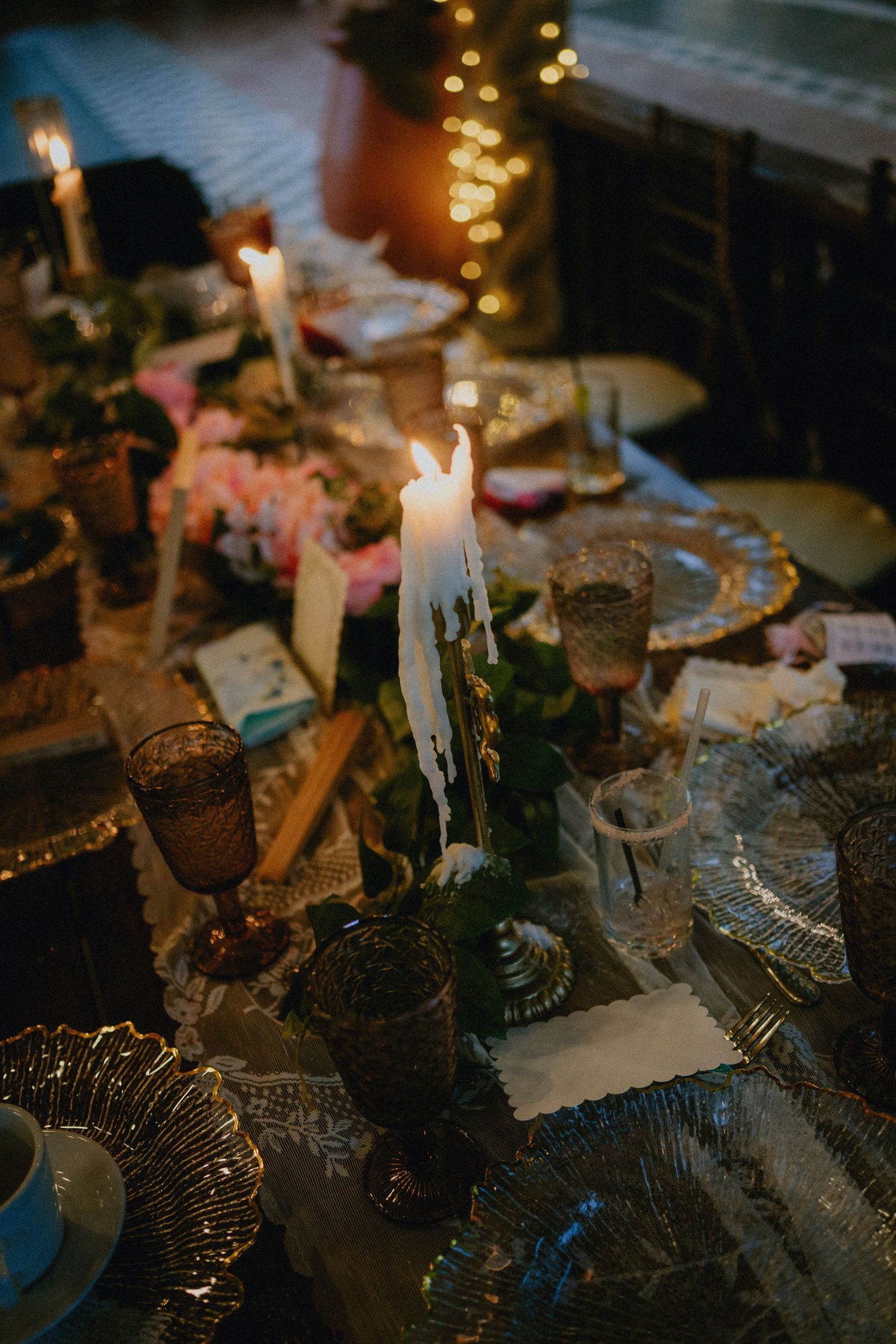Elegant candlelit dinner table with dripping white candles, vintage glassware, gold chargers, and floral centerpieces.