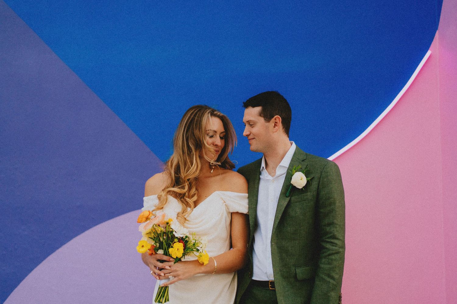 Bride and groom pose before colorful blue and pink mural, bride holding vibrant yellow bouquet in white off-shoulder gown.