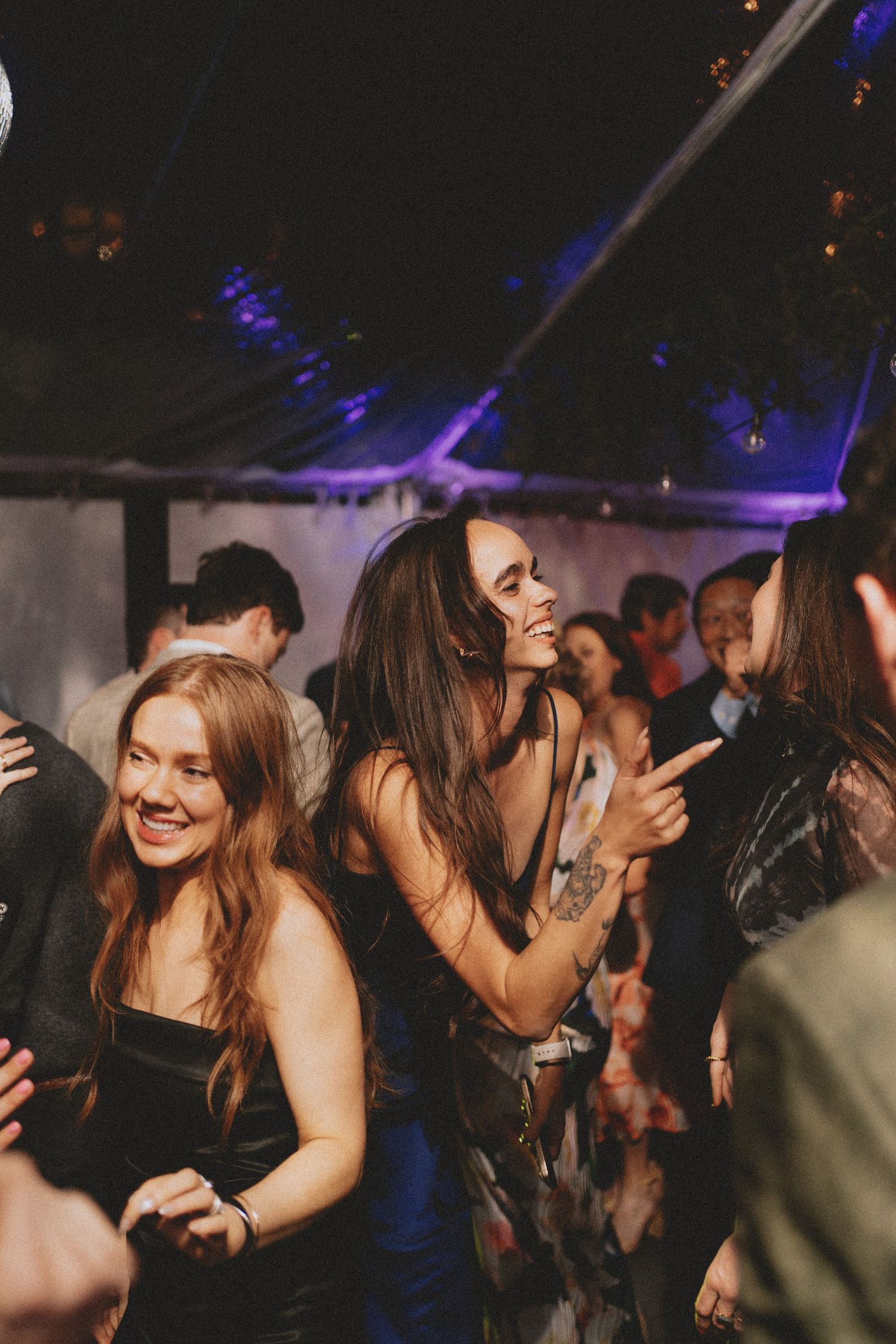 Guests dancing joyfully at wedding reception under a tented venue with purple lighting.