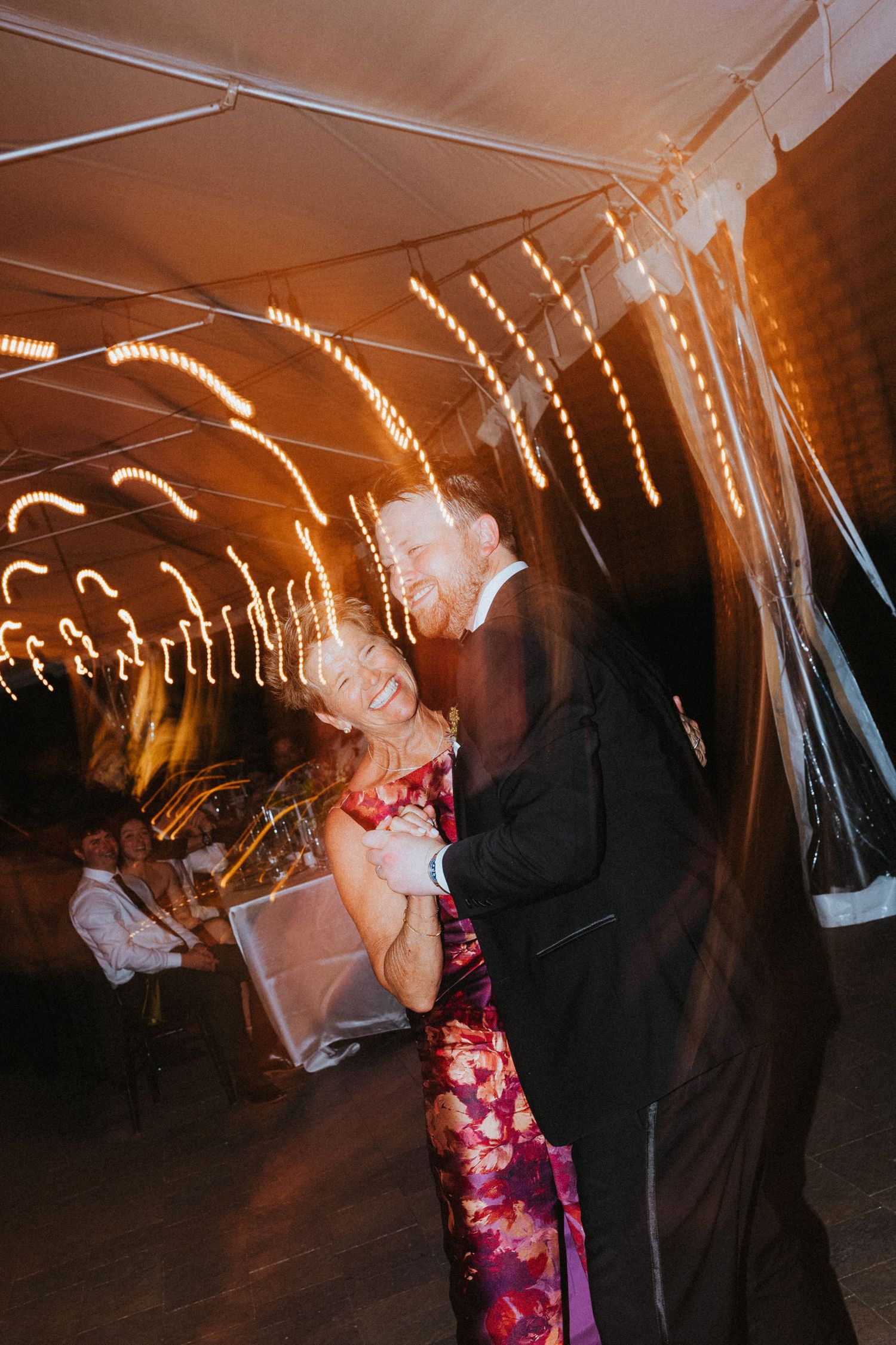 Couple shares a joyful dance under warm string lights with motion blur at a wedding reception.