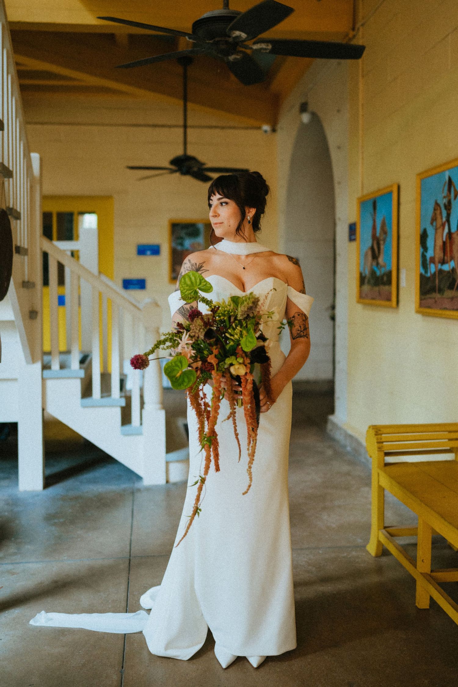 Bride in white off-shoulder gown holding lush cascading bouquet stands in yellow-accented venue hallway.