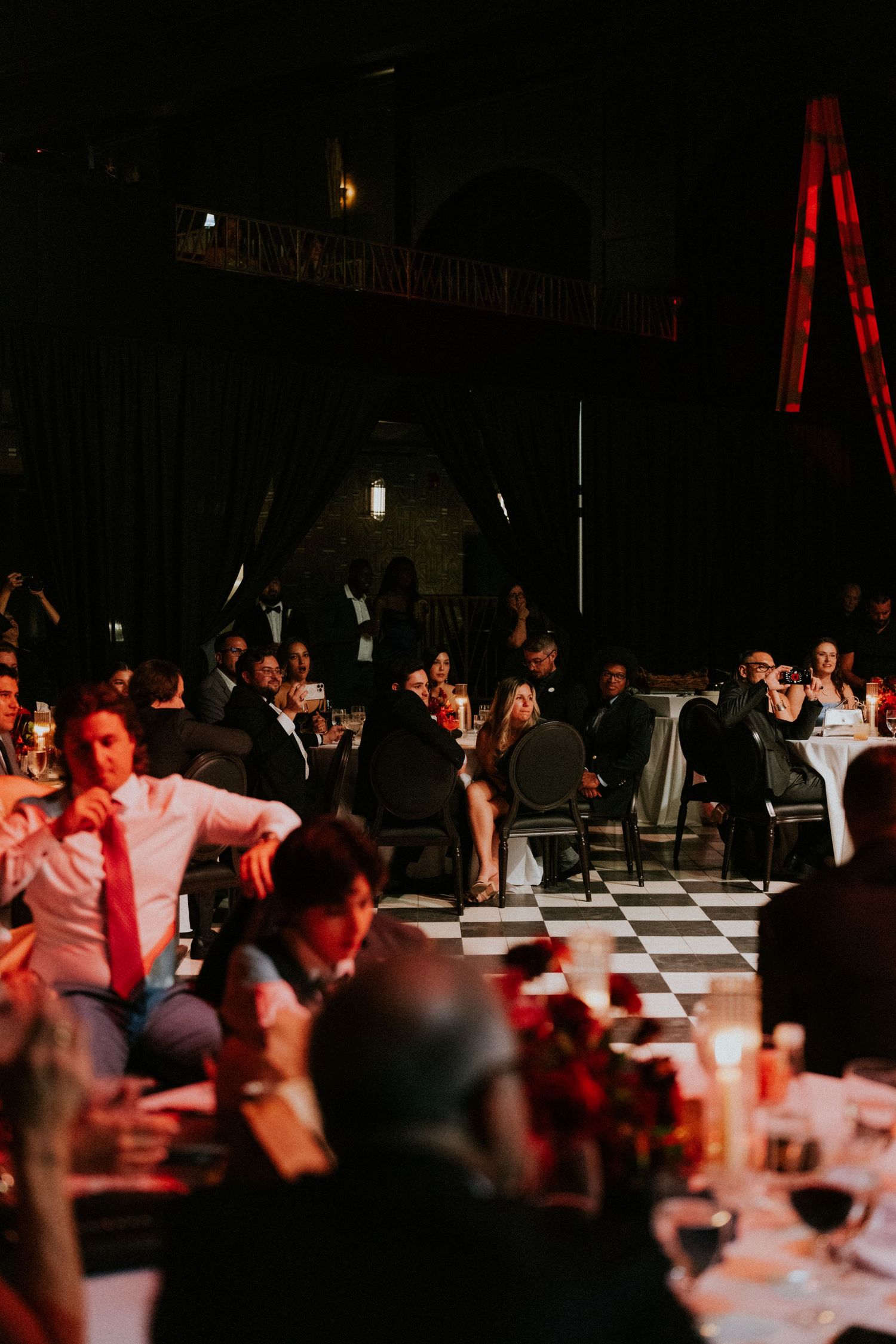 Elegant gala dinner reception with guests seated at candlelit tables on a checkered dance floor in a grand dark venue.