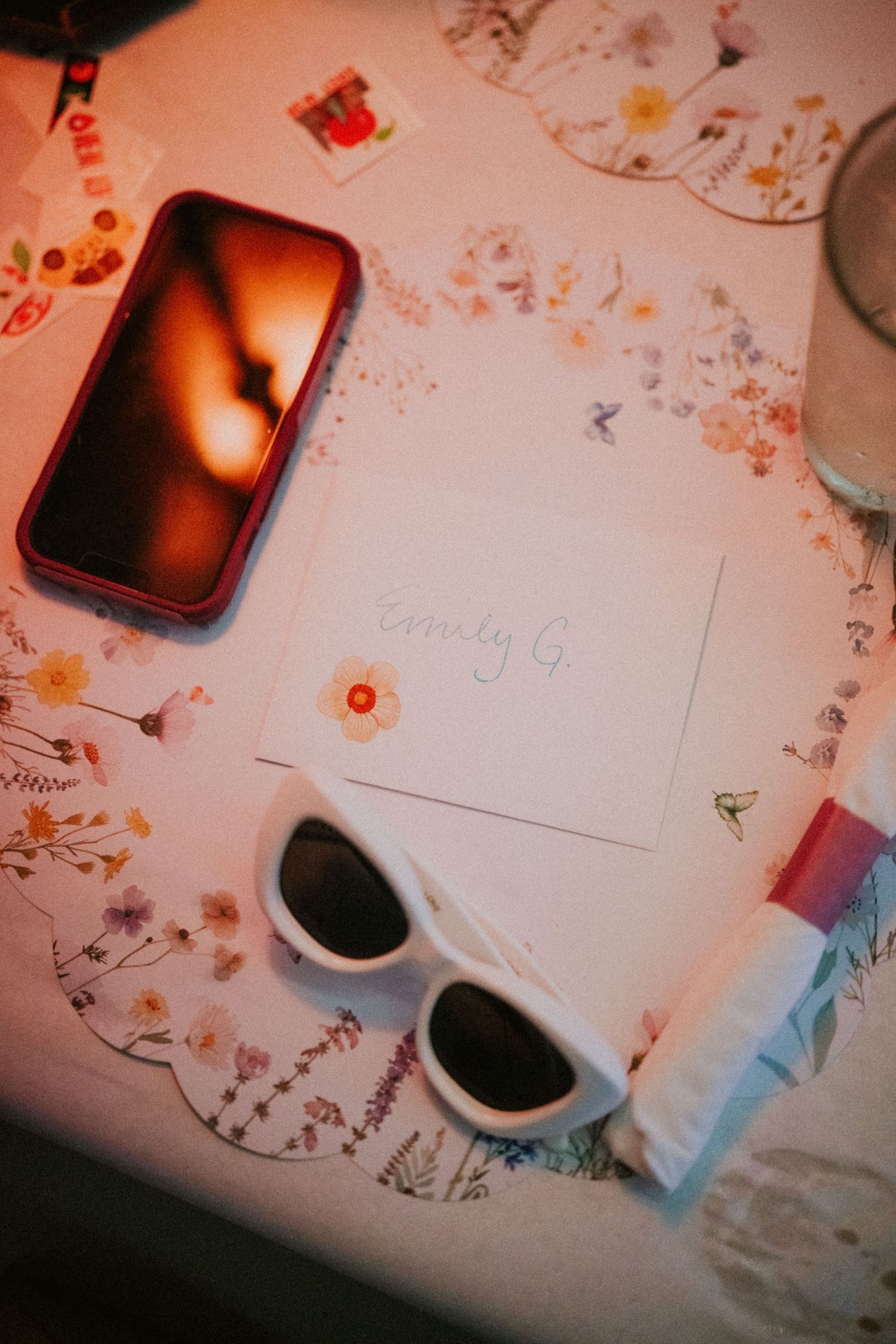 Flat lay of white sunglasses, a name card reading Emily G., a smartphone, and floral decorations on a white table.