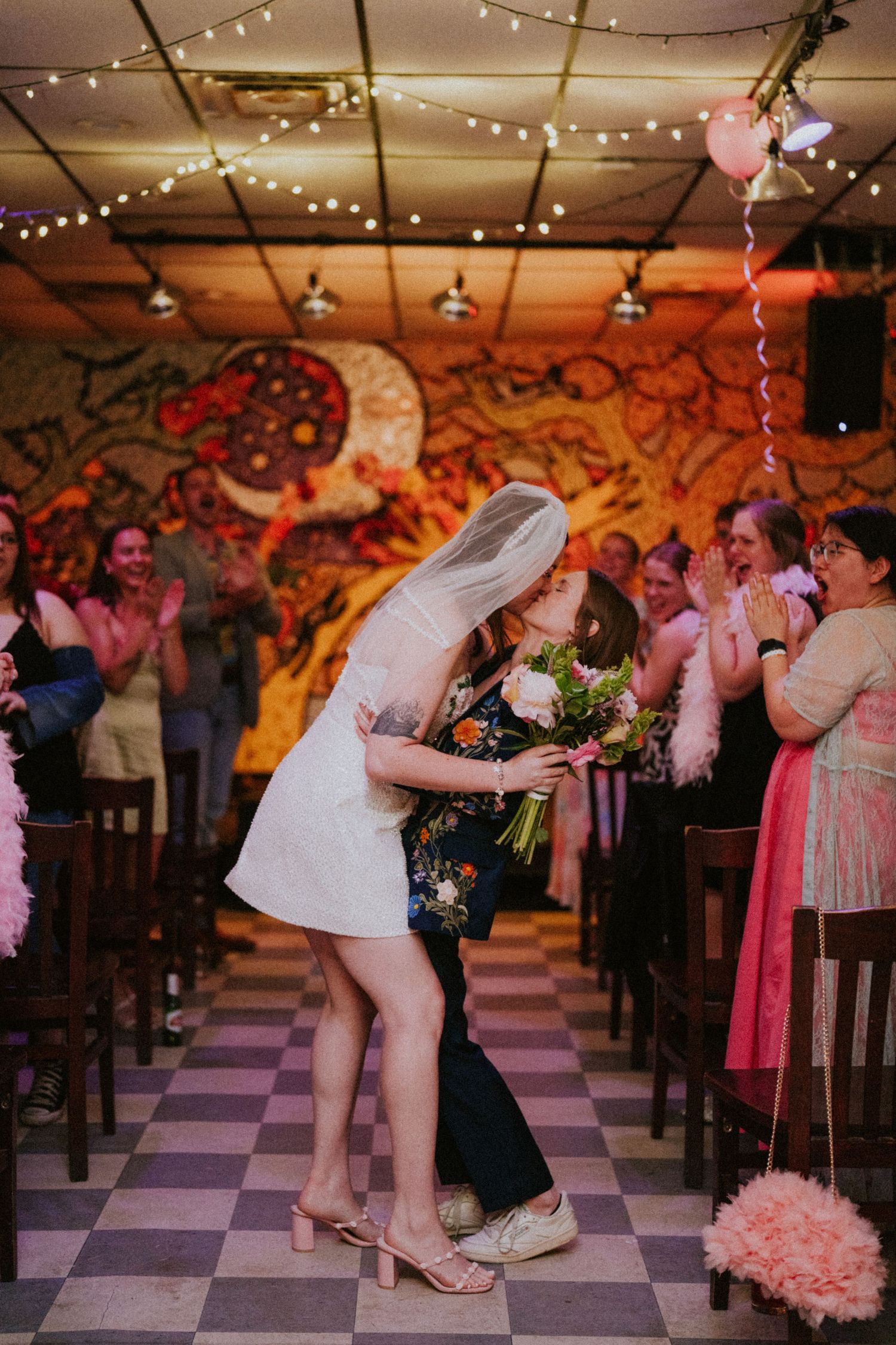 Two brides share a first kiss in the aisle of a decorated venue with string lights and a colorful mural.