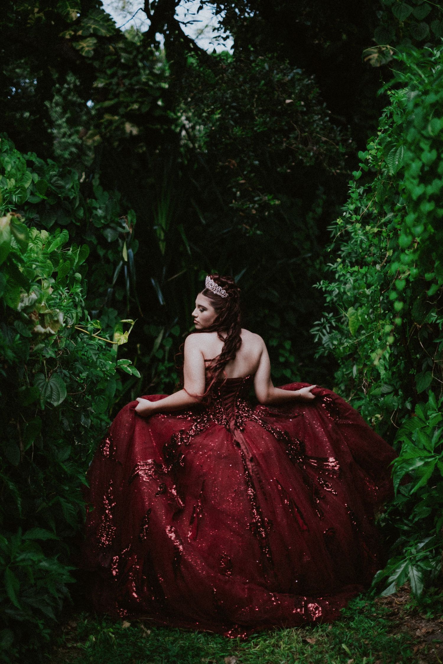 A figure in a flowing burgundy sequined gown poses dramatically against lush green foliage in an enchanted forest setting.