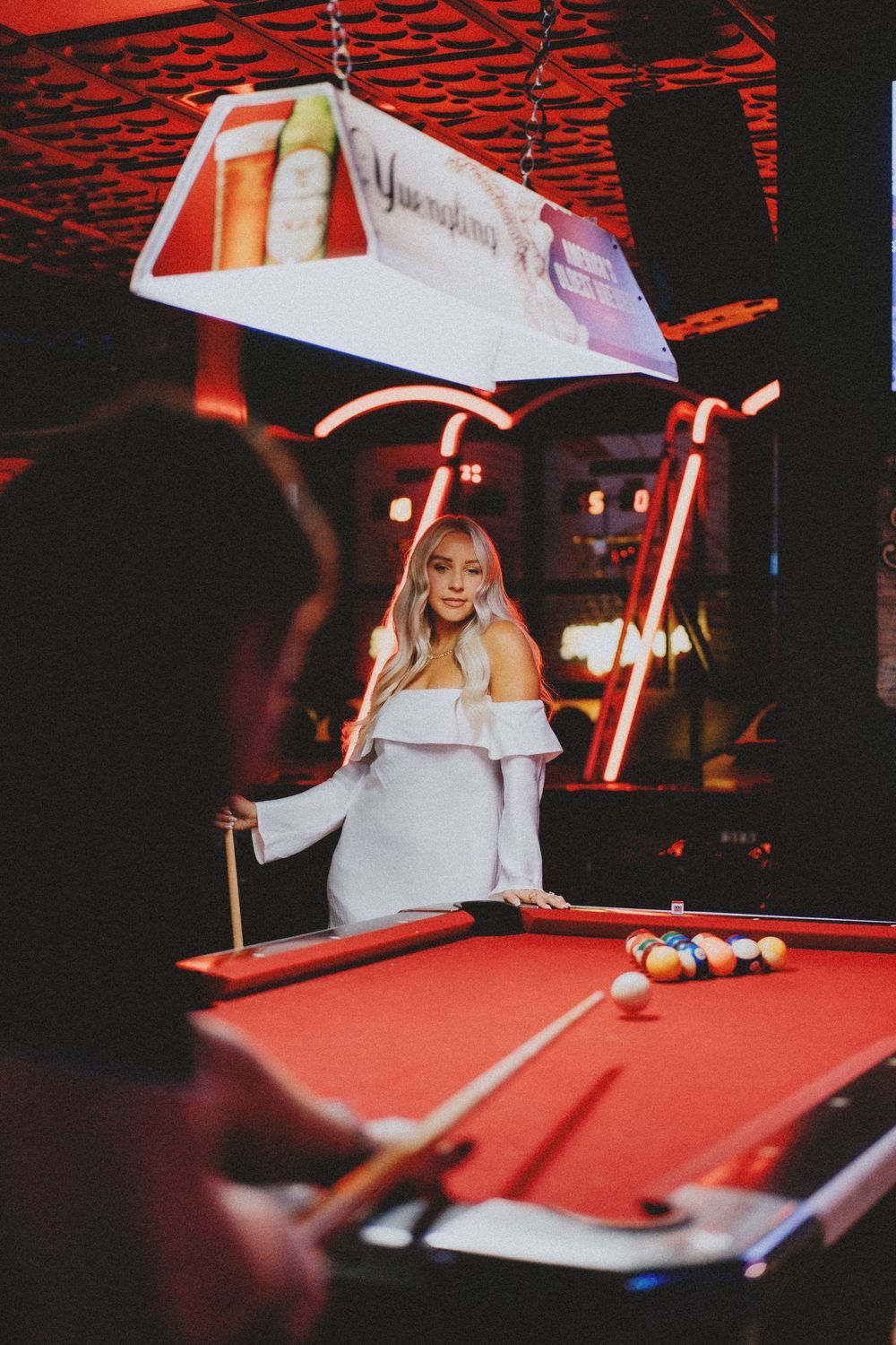 Person in white playing pool at a dimly lit bar with red neon lighting.