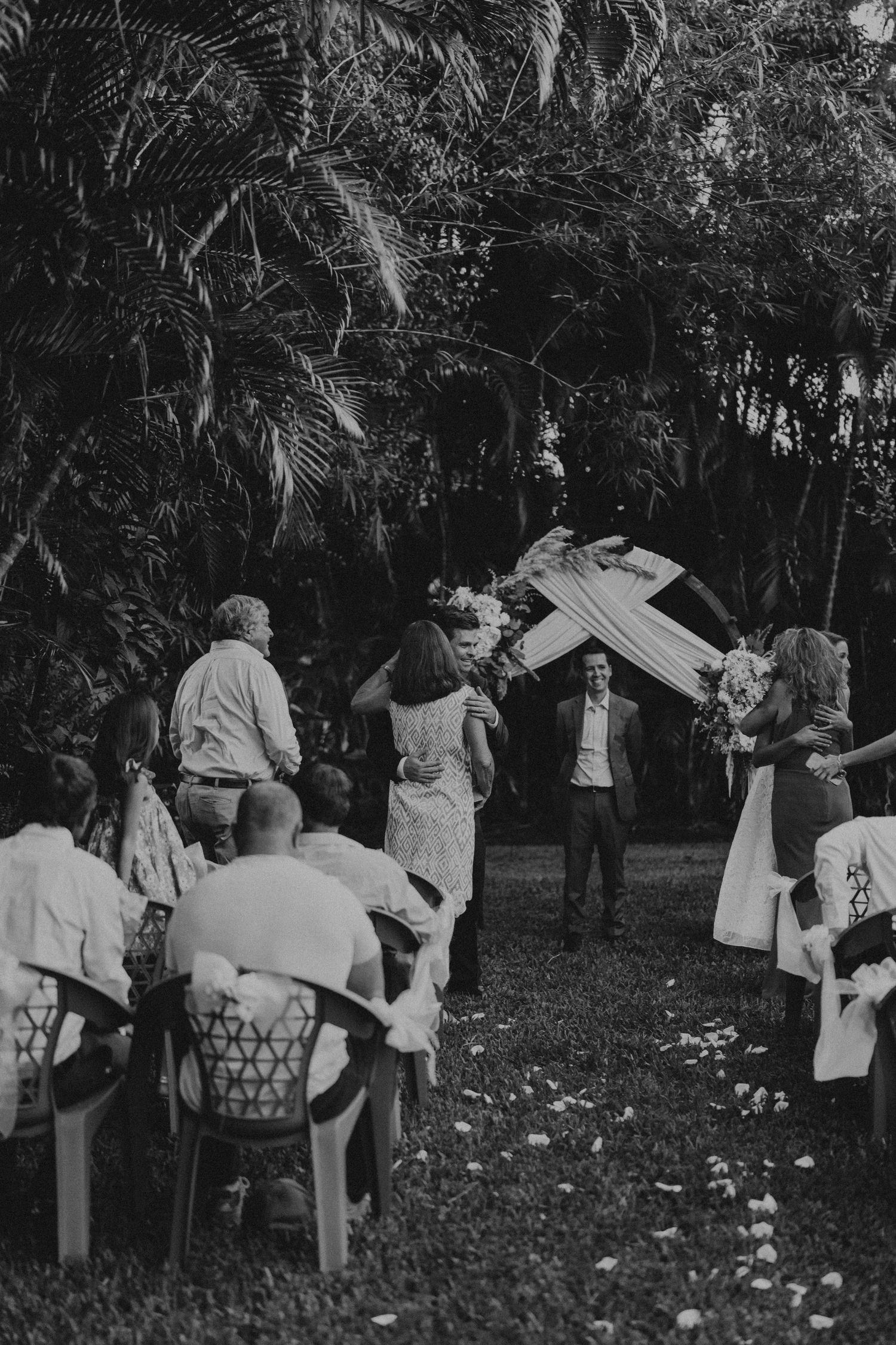 A black and white photo of an outdoor wedding ceremony under palm trees with guests seated in chairs.