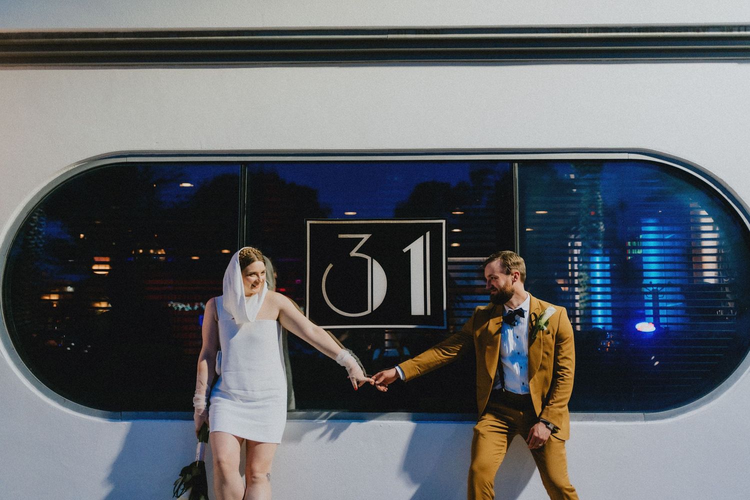 Bride and groom in wedding attire hold hands walking past art deco building with number 31 at night.