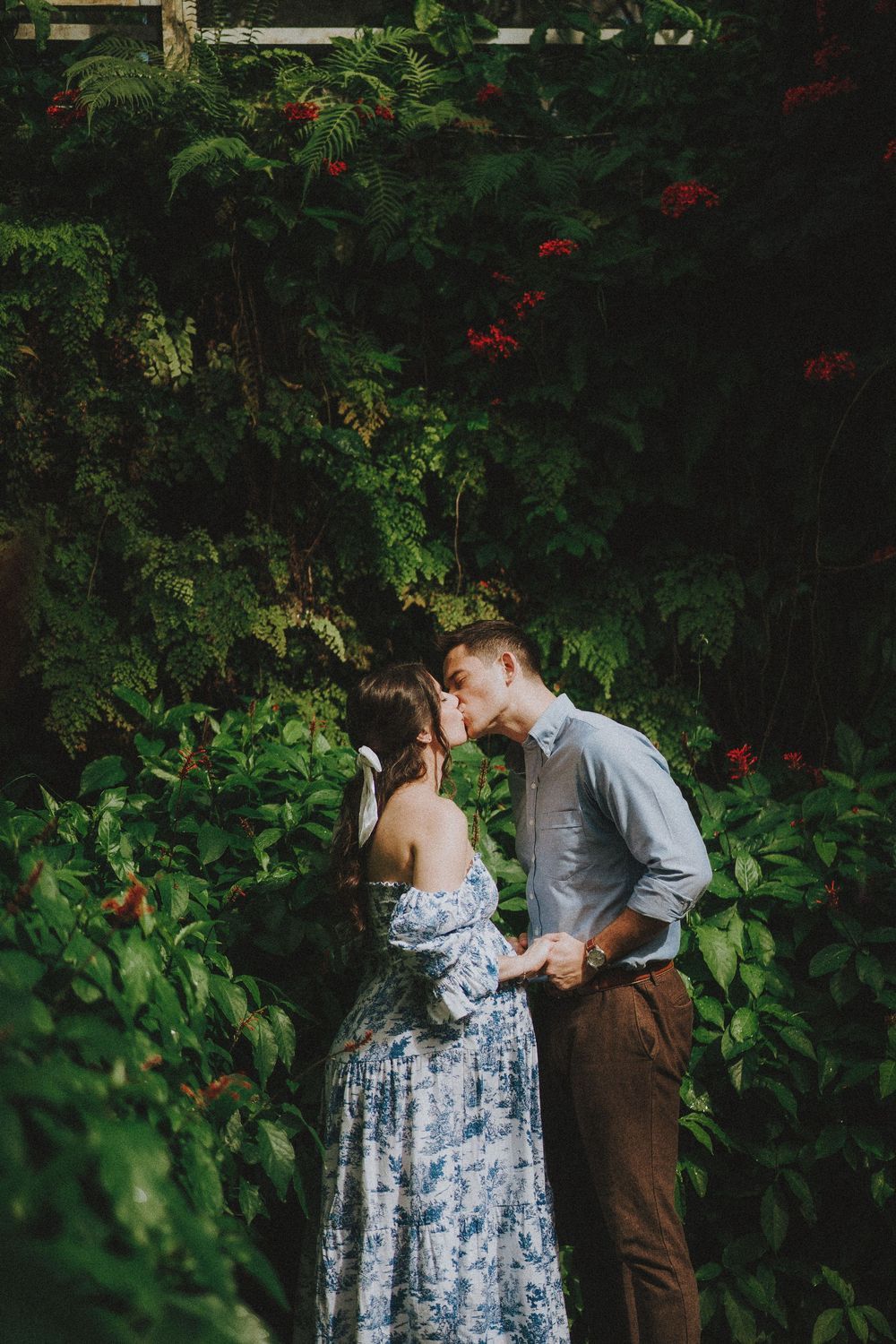 A couple shares an intimate moment in a lush garden setting with vibrant greenery and red flowers.