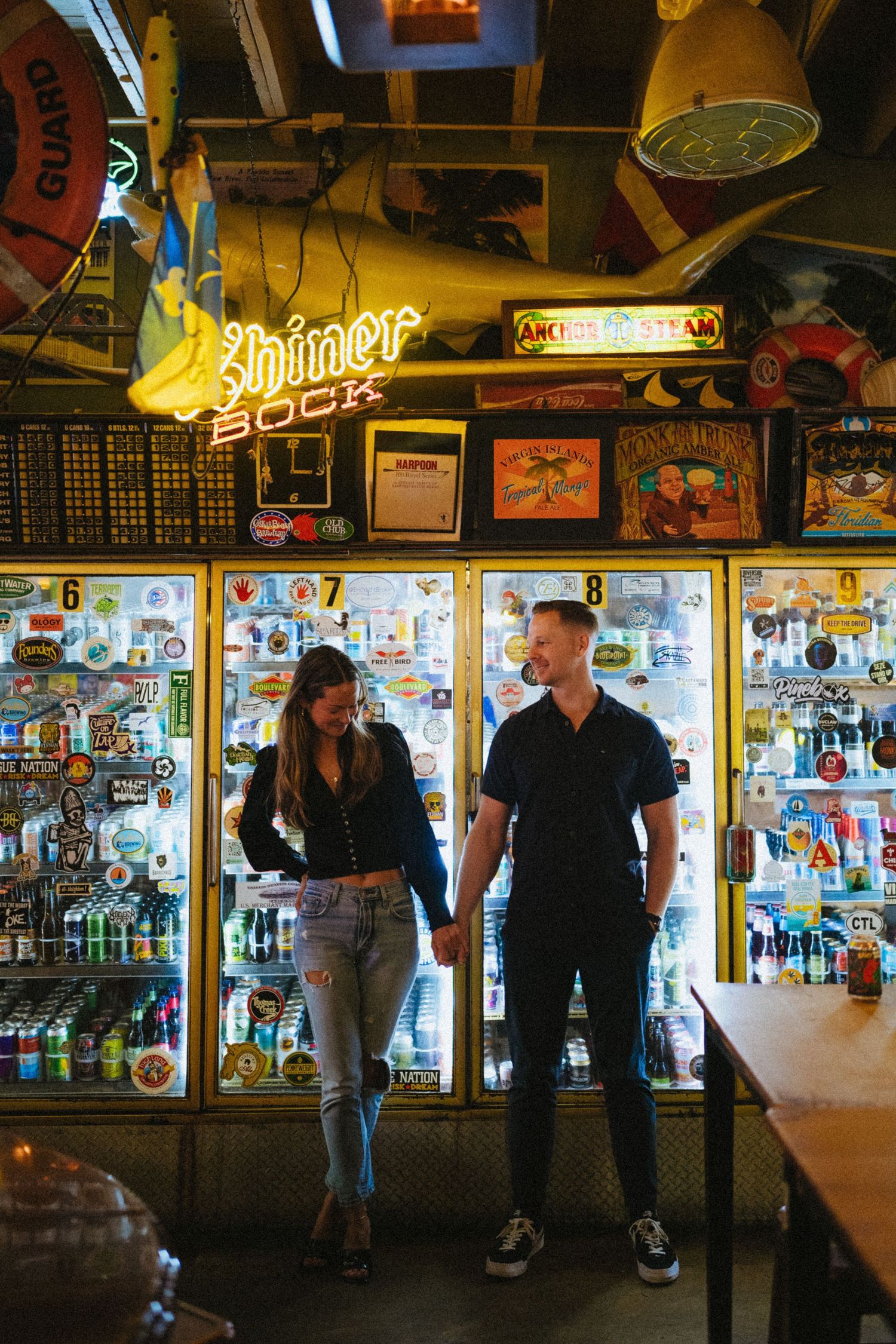 Couple holding hands in front of illuminated beer fridges under a Shiner Bock neon sign in a bar.