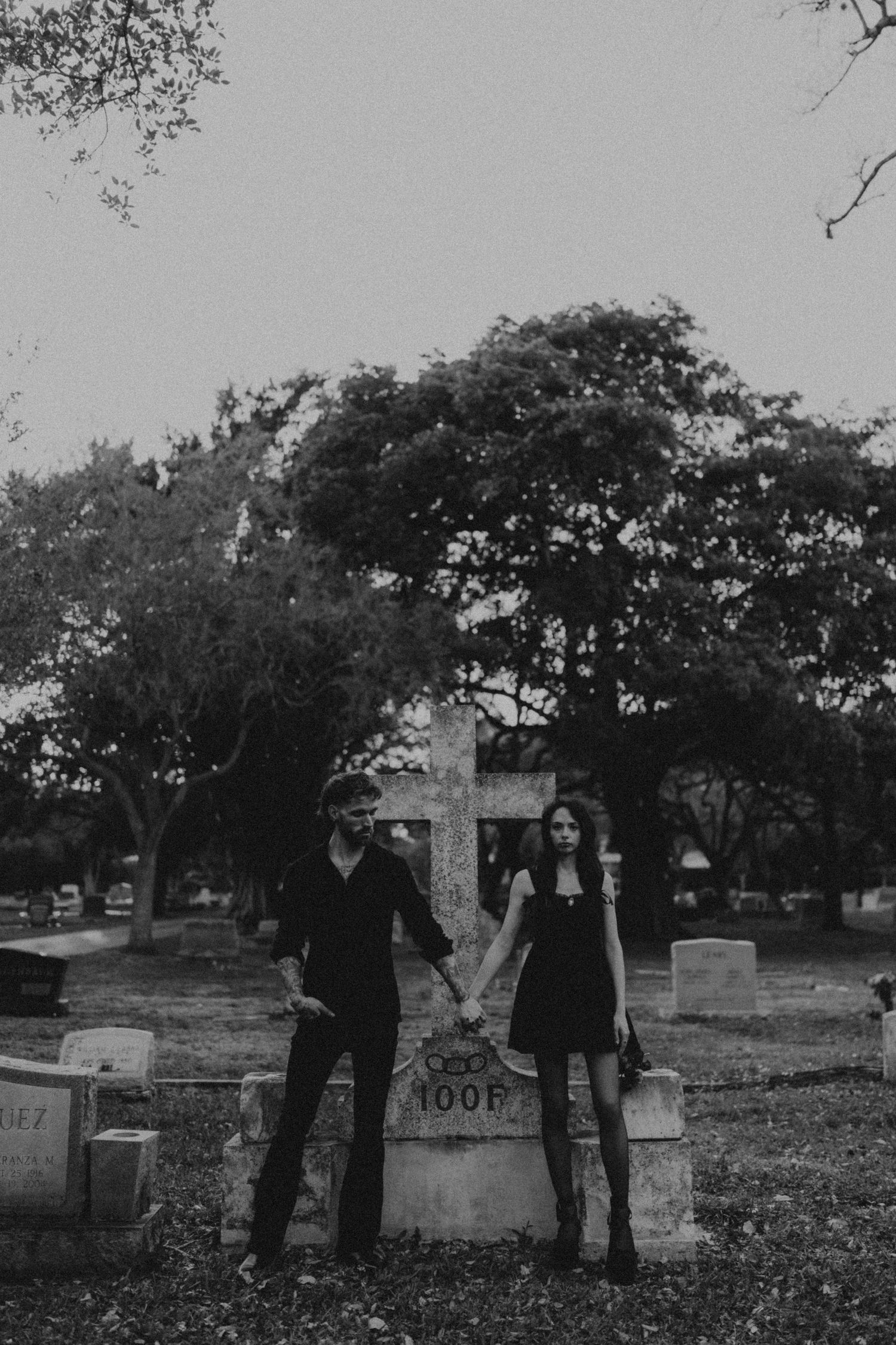 Couple in dark attire posing by a cross gravestone in a cemetery, black and white gothic photo.