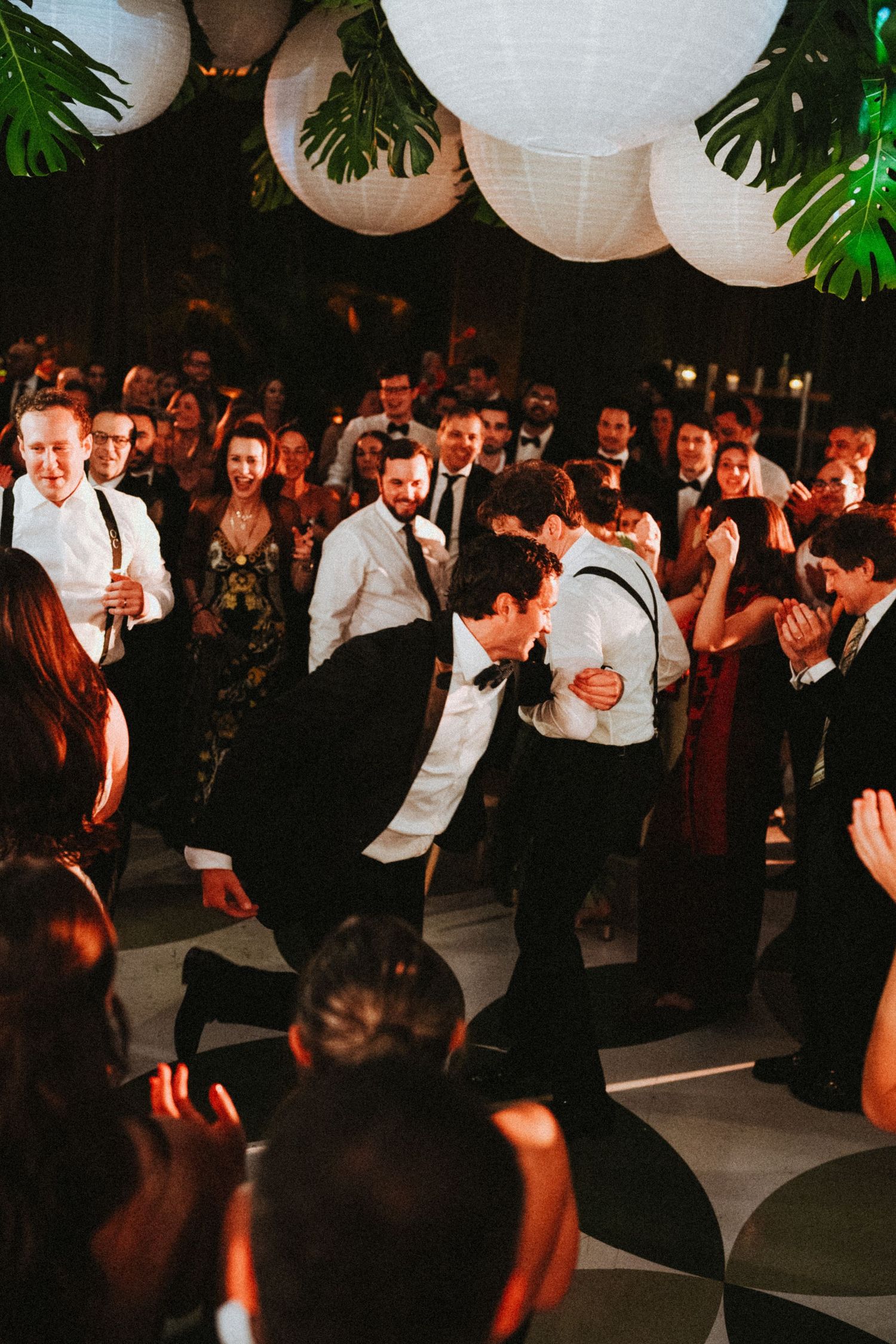 Two grooms share a joyful dance moment surrounded by cheering wedding guests under white paper lantern decorations.