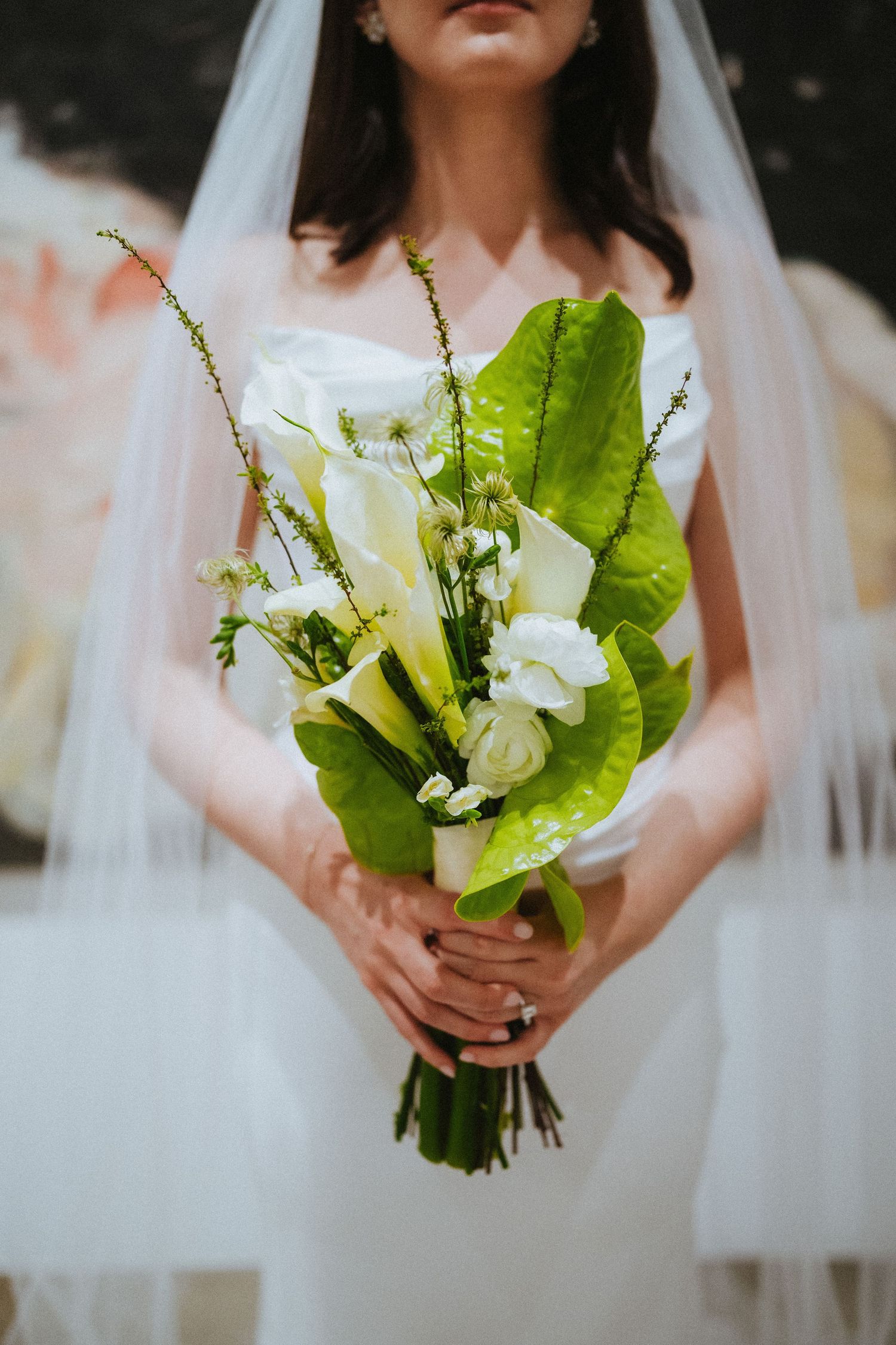 Bride in white gown and veil holding elegant bouquet of white calla lilies and green tropical leaves.