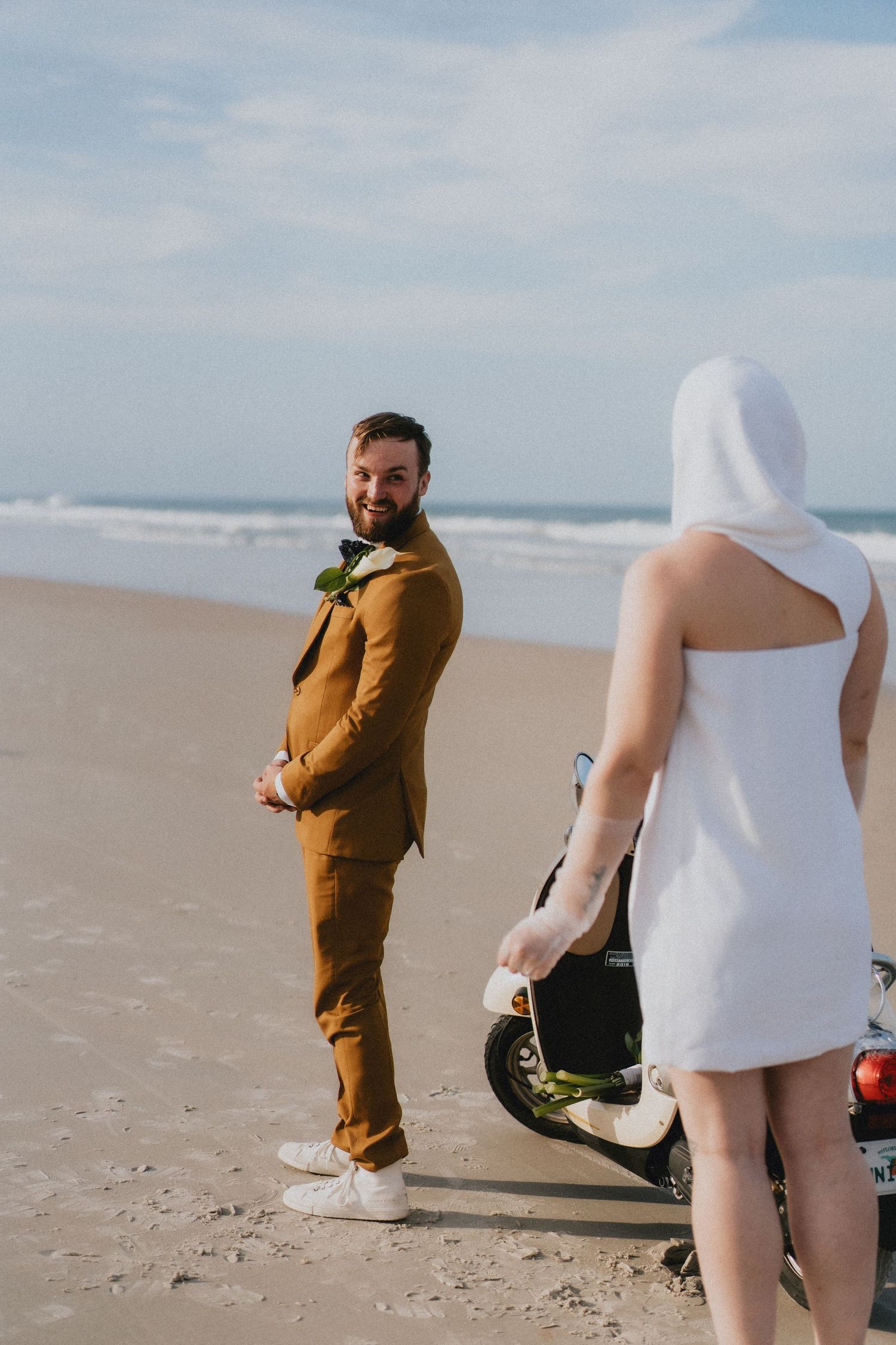 Smiling groom in mustard suit on beach looks back at bride in white hooded dress near scooter.