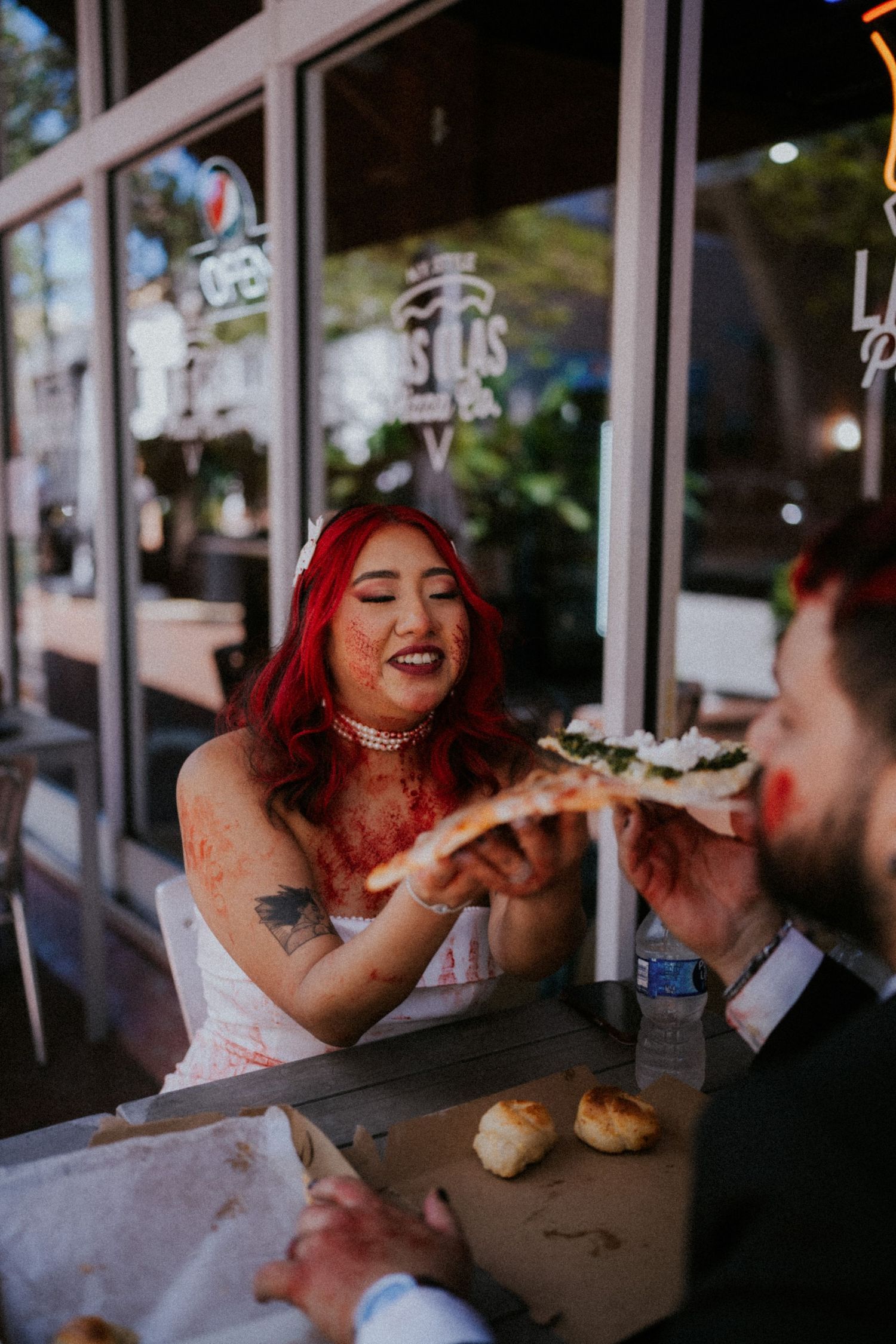 Red-haired bride in white dress shares pizza slice with groom outside restaurant on wedding day.