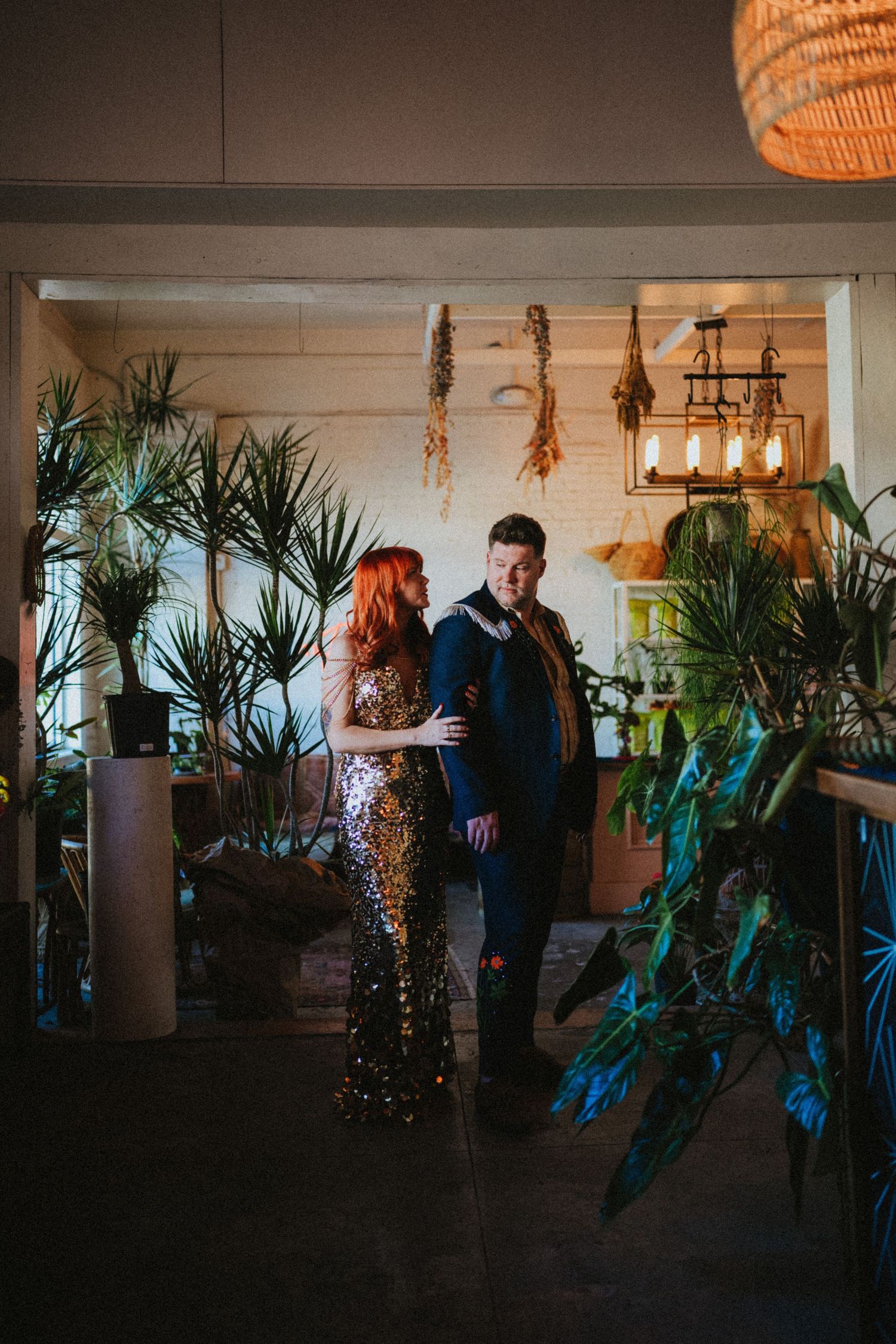Couple in elegant attire stand among lush tropical plants in a warmly lit venue, woman in gold sequin gown.