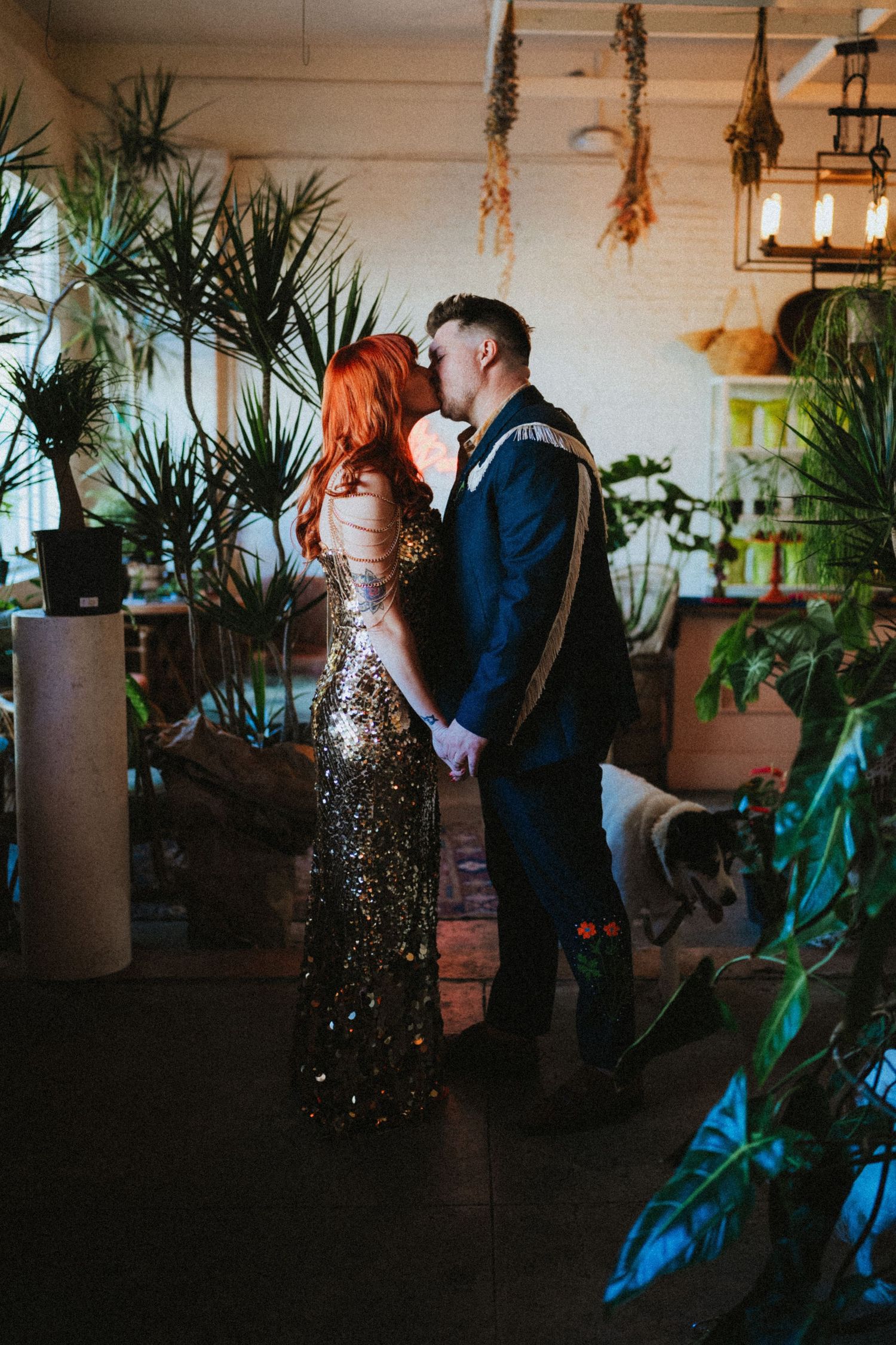 Bride in gold sequin gown and groom in navy suit share a kiss surrounded by lush indoor plants.