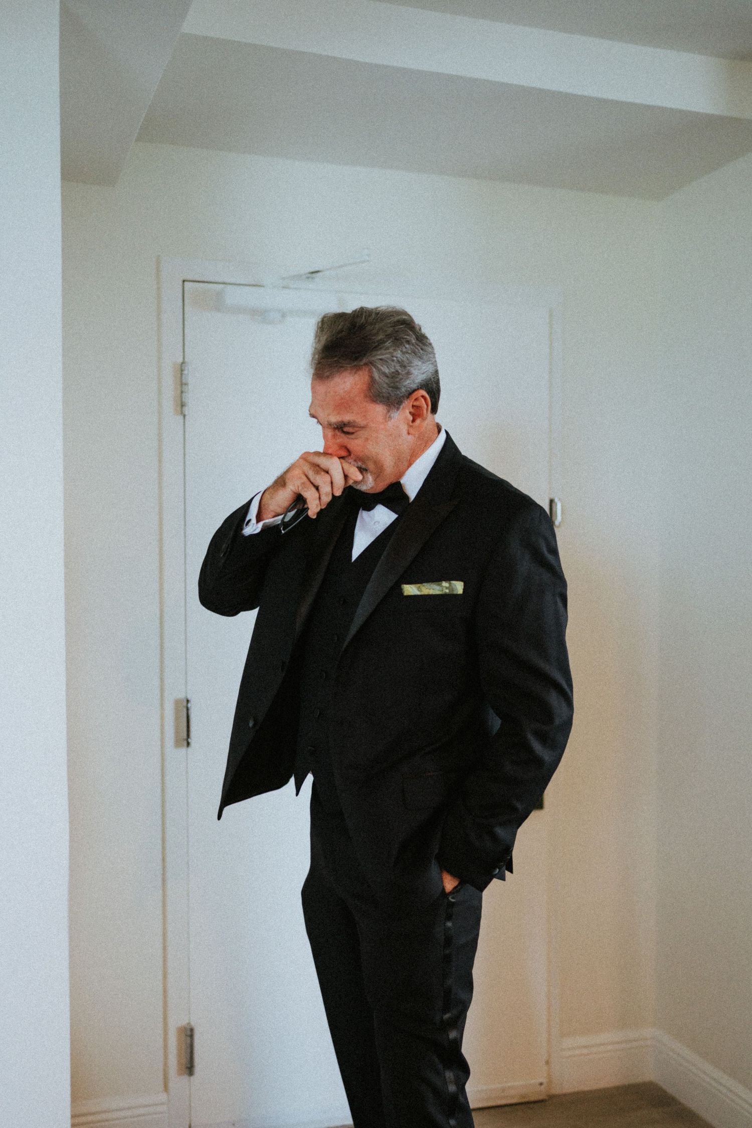 Emotional groom in black tuxedo with bow tie and yellow pocket square, covering mouth while looking down.