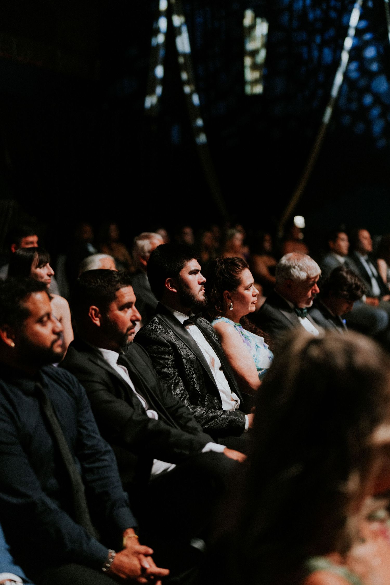 Elegantly dressed wedding guests seated in rows, attentively watching a ceremony in a dimly lit outdoor venue.