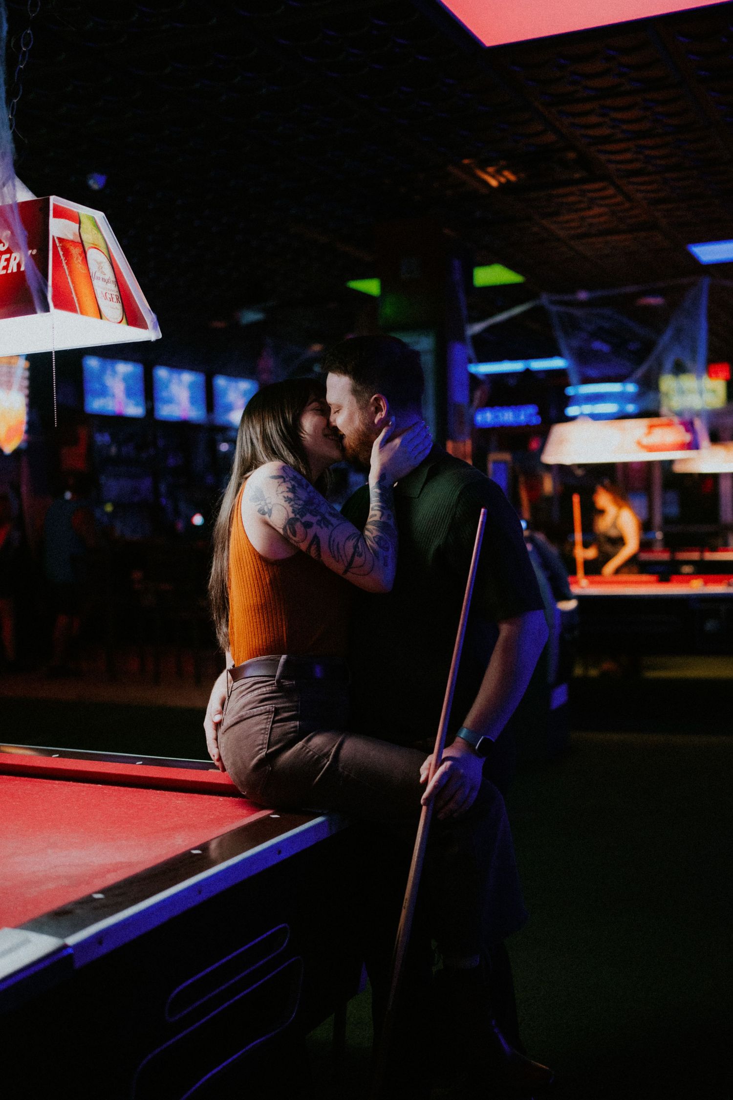 Couple sharing a romantic kiss leaning on a pool table in a dimly lit bar with neon lights.