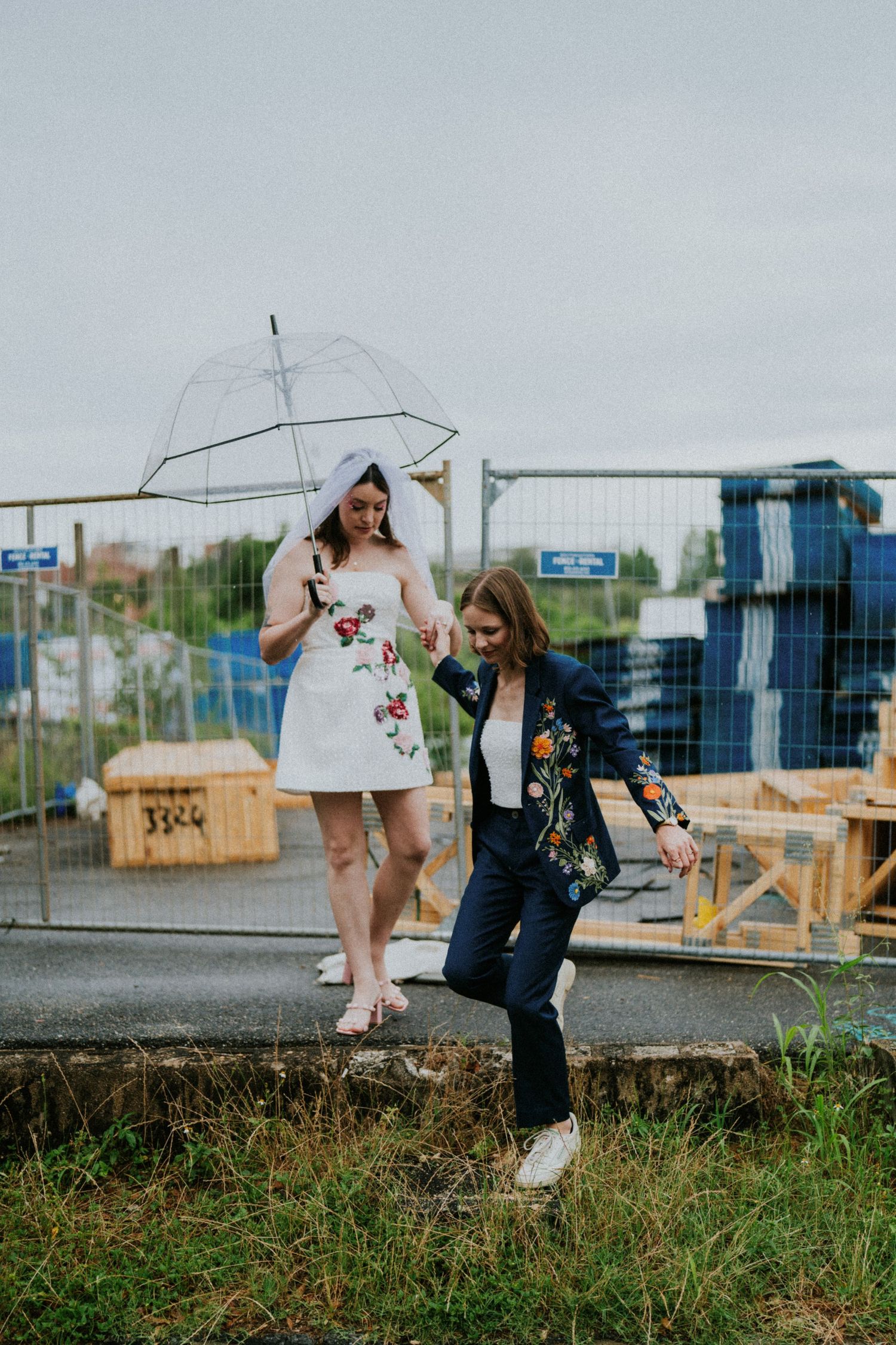 Two brides walk hand-in-hand in the rain, one holding a clear umbrella, wearing a floral dress and navy embroidered blazer.