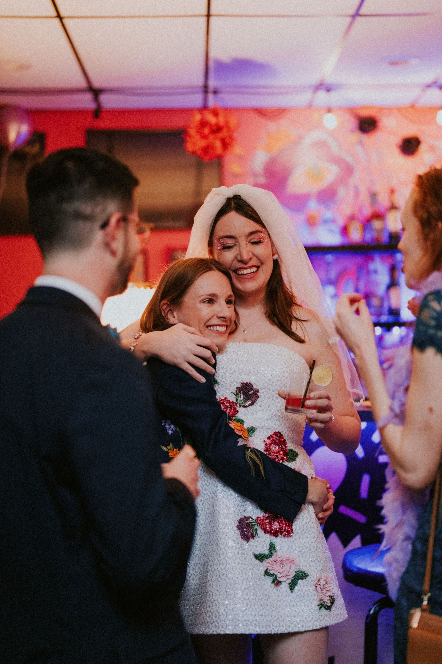 Bride in white floral mini dress and veil hugs a guest while laughing at a colorful wedding reception.