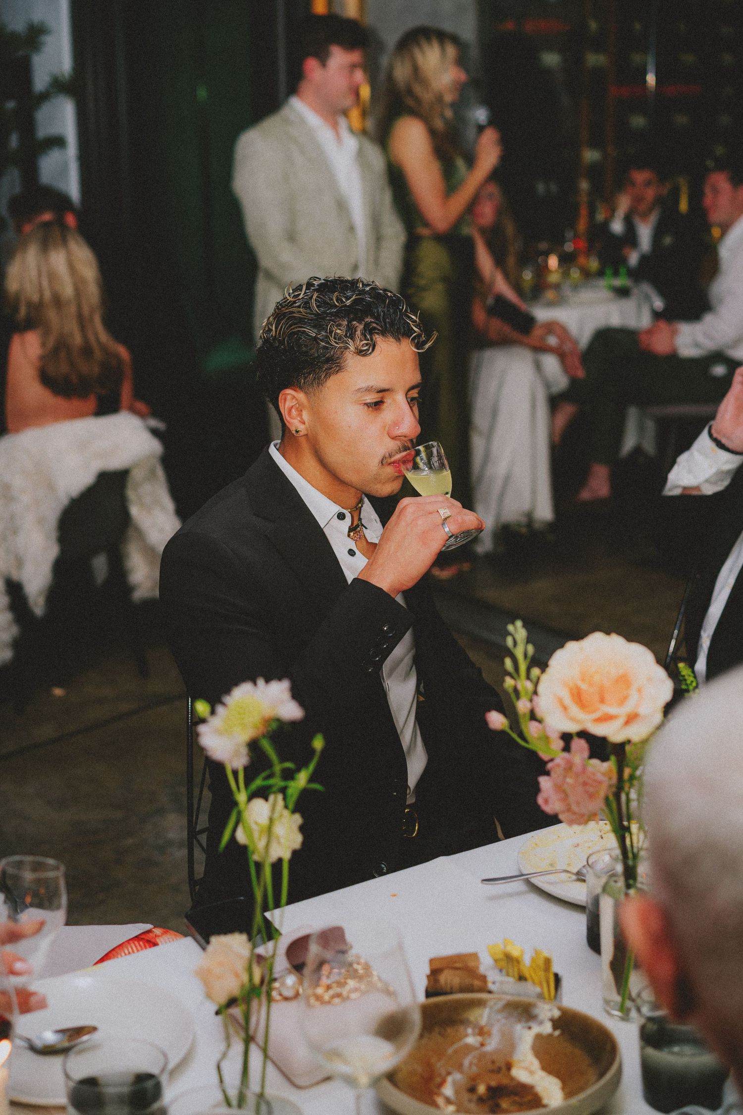 A young man in a black suit sips champagne at an elegant wedding reception dinner table with floral centerpieces.