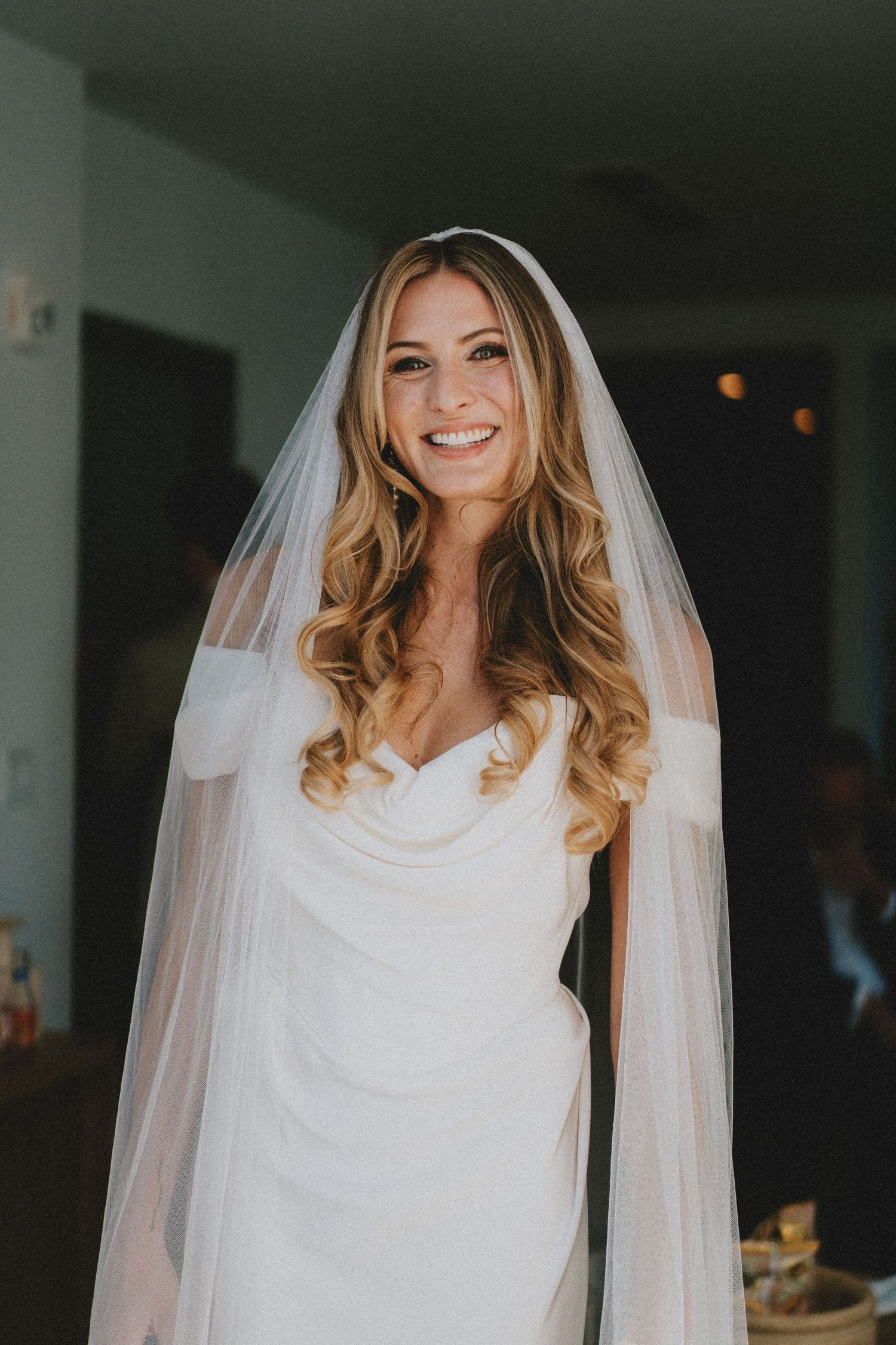 Smiling bride in white off-shoulder gown and long flowing veil with wavy blonde hair on wedding day.
