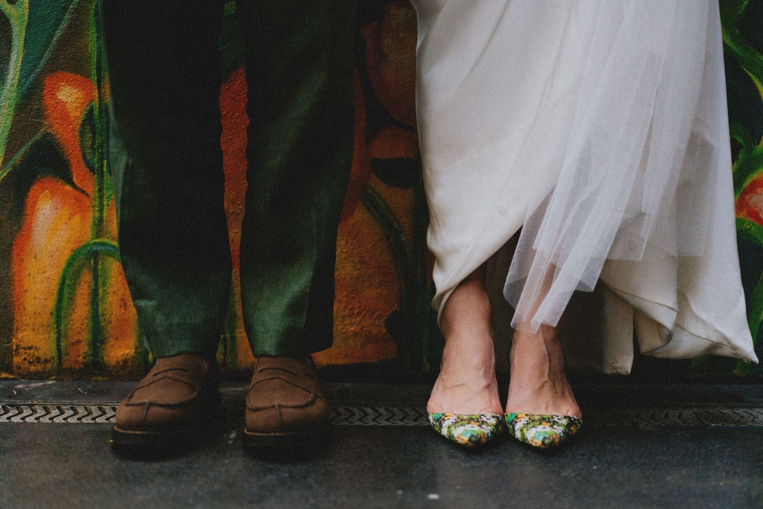 Close-up of wedding couple's feet, groom in brown loafers and bride in floral embellished heels on dark floor.